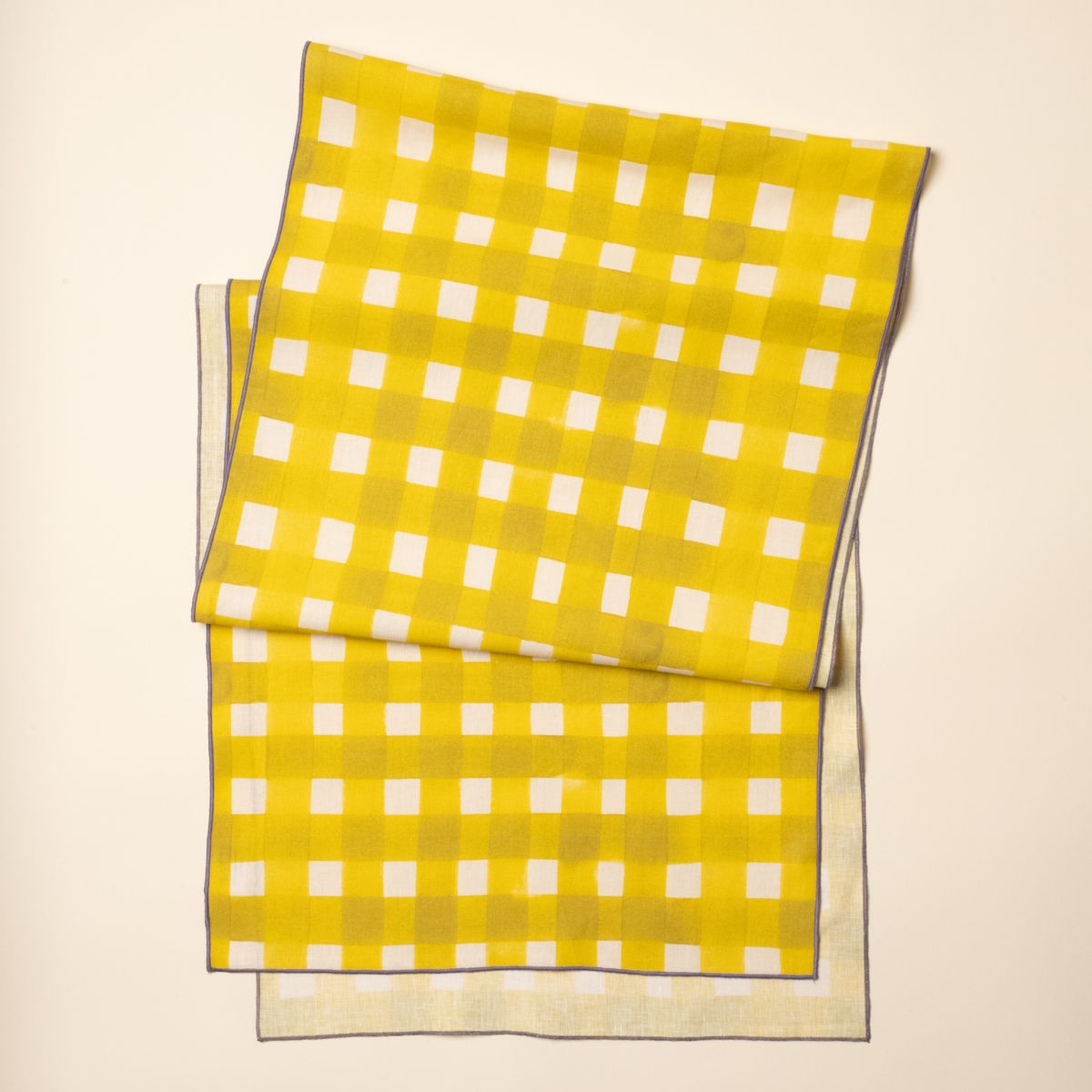 A table runner folded laying flat featuring a bright yellow crosshatch pattern with serged purple edges.