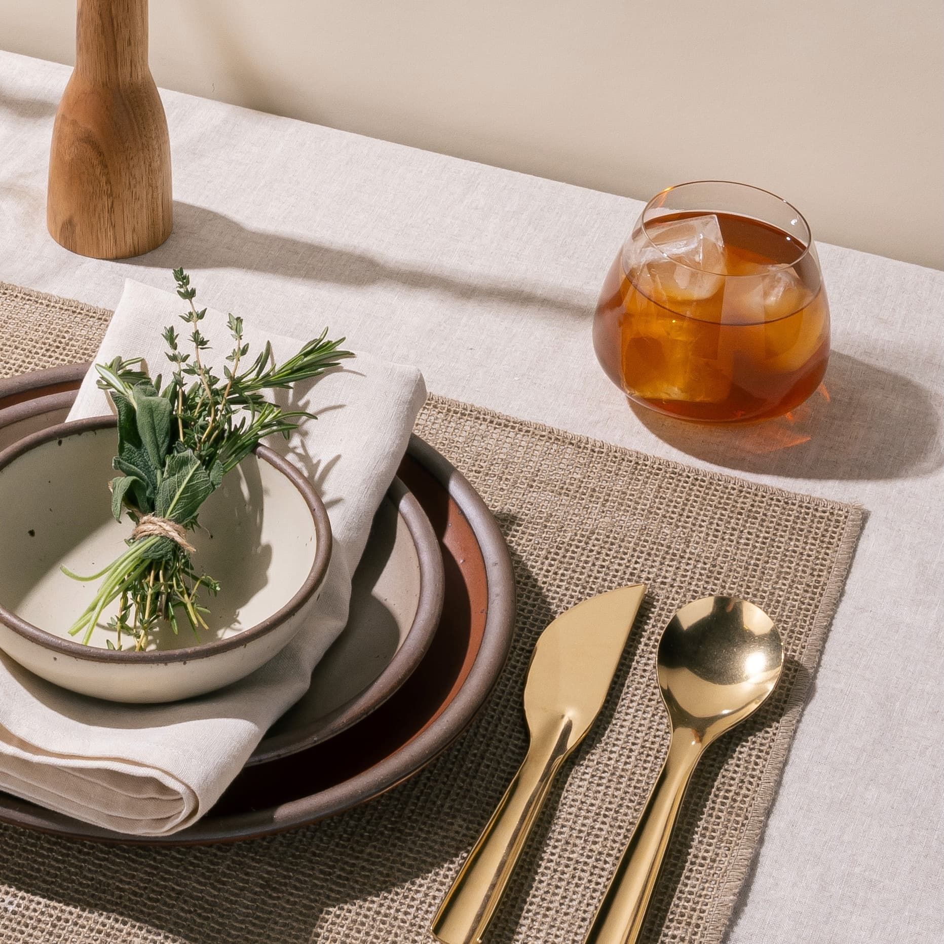 A clear glass whiskey snifter with a rounded base with a cocktail inside sits on a table near a place setting. The place setting features ceramic dinnerware, tied herbs, and brass flatware 