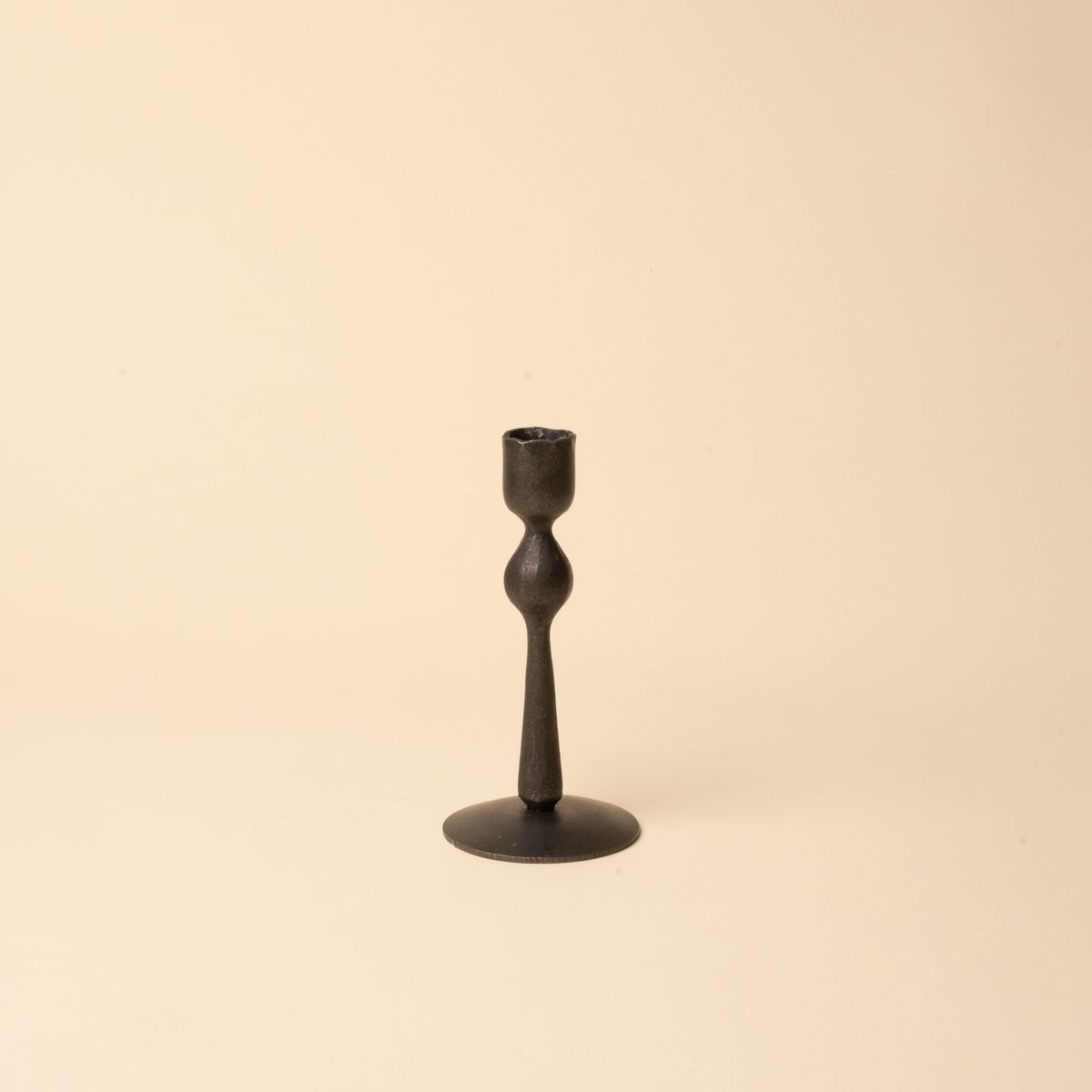 Short minimal black forged iron candlestick with a sculptural shape on a neutral beige background.