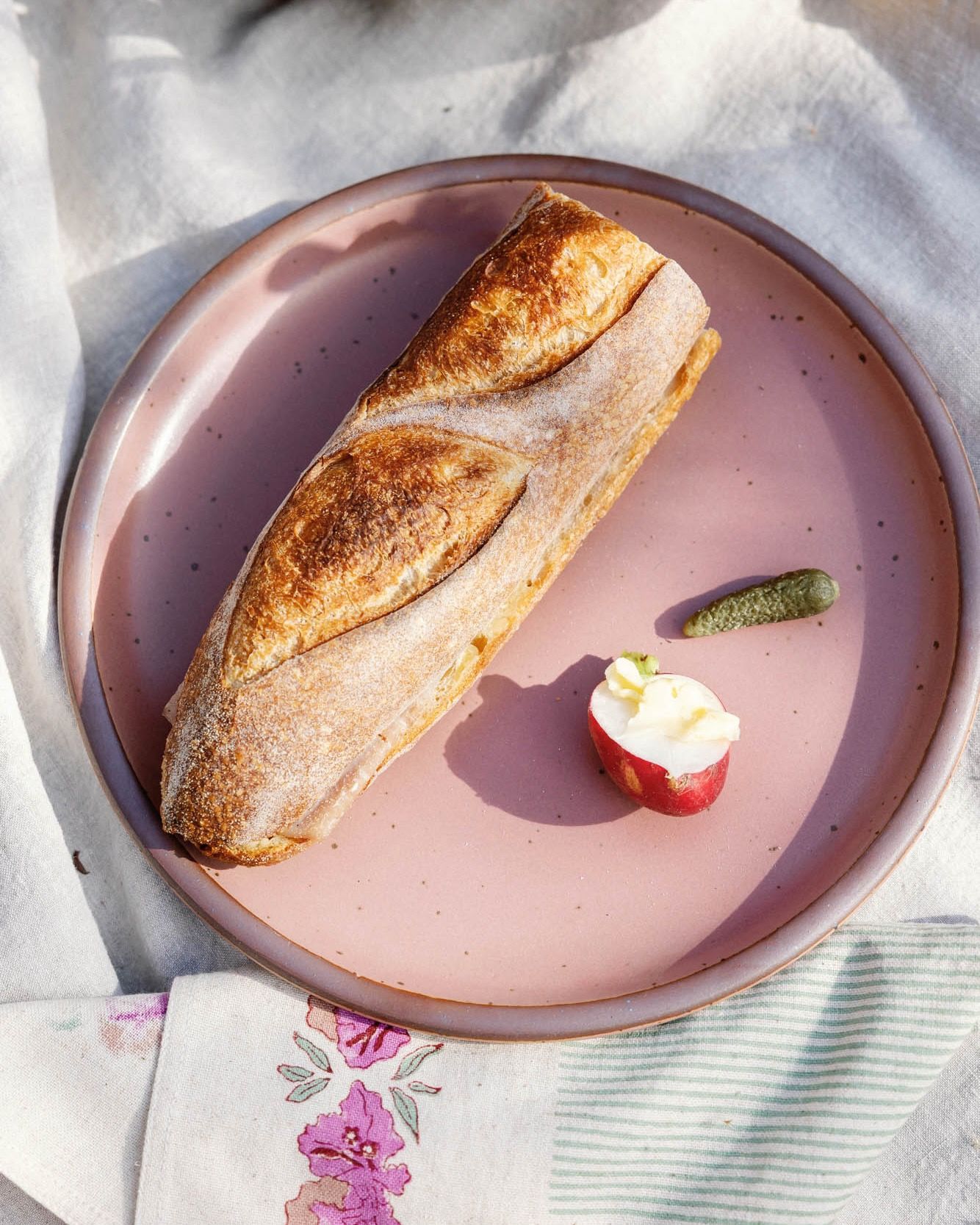 Pink ceramic plate with a baguette sandwich, a radish with butter, and a small pickle on a linen tablecloth.