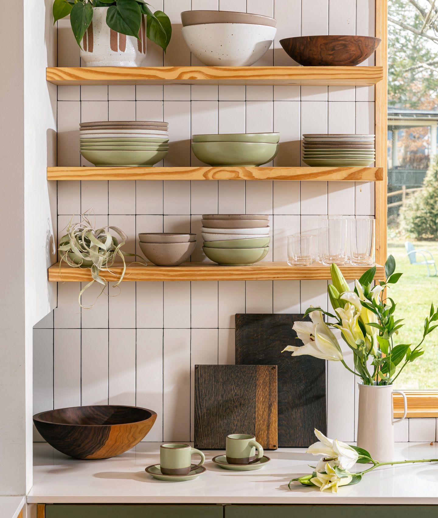 In a kitchen setting, there are floating shelves above a countertop filled with stacks of bowls and plates, simple drinking glasses, and an air plant. On the countertop is small mugs sitting on saucers, a large walnut bowl, 2 large dark cutting boards, and a water pitcher filled with flowers.