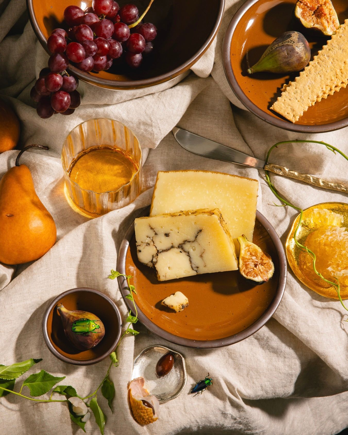 Glossy caramel brown plate with truffle cheese, figs, grapes, crackers, honeycomb, and a glass of amber drink on linen.
