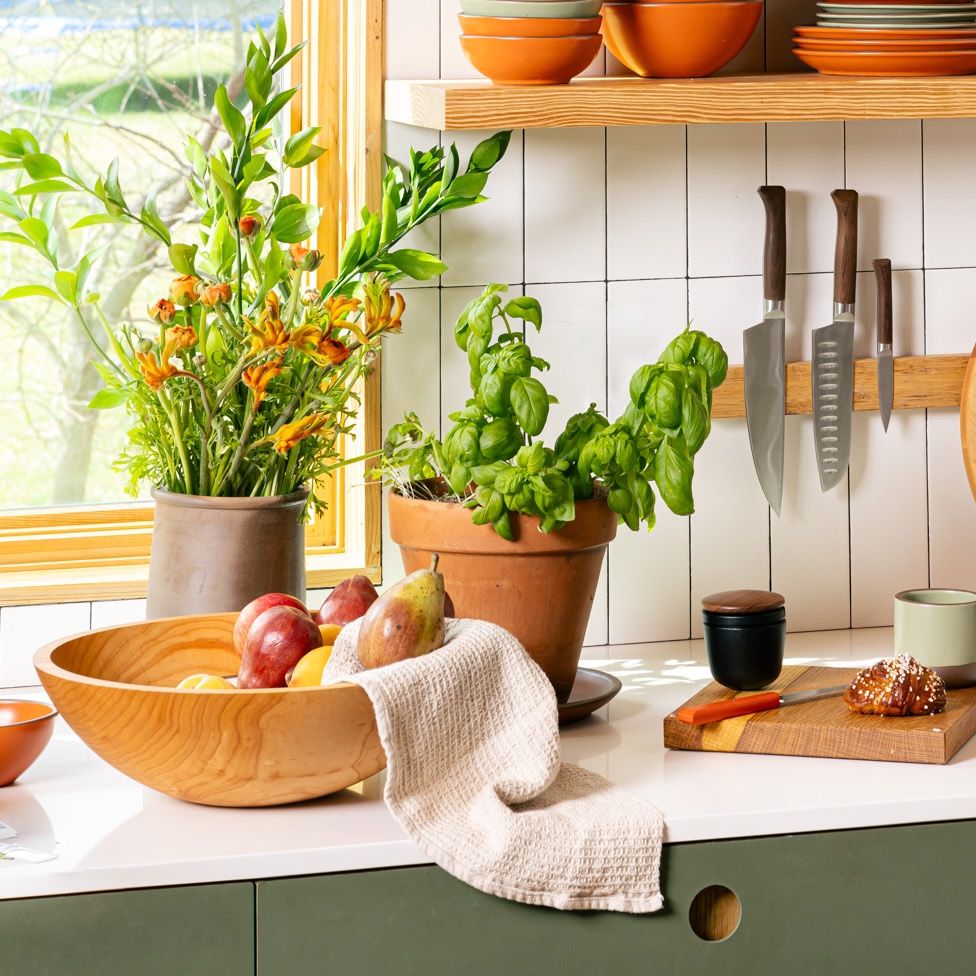 On a kitchen countertop sits a large maple bowl filled with fruit, a natural waffle kitchen towel, potted plants, and a knife block on the wall with a cutting board.