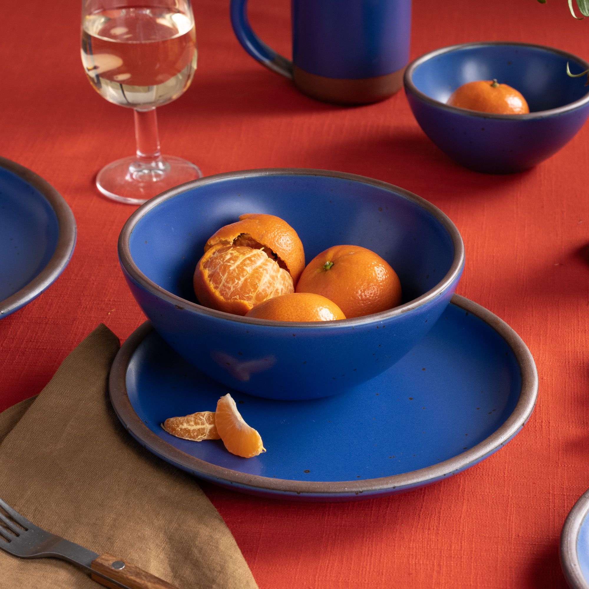 Blue ceramic bowl and plate with clementines on a red tablecloth beside a glass of white wine and brown napkin.