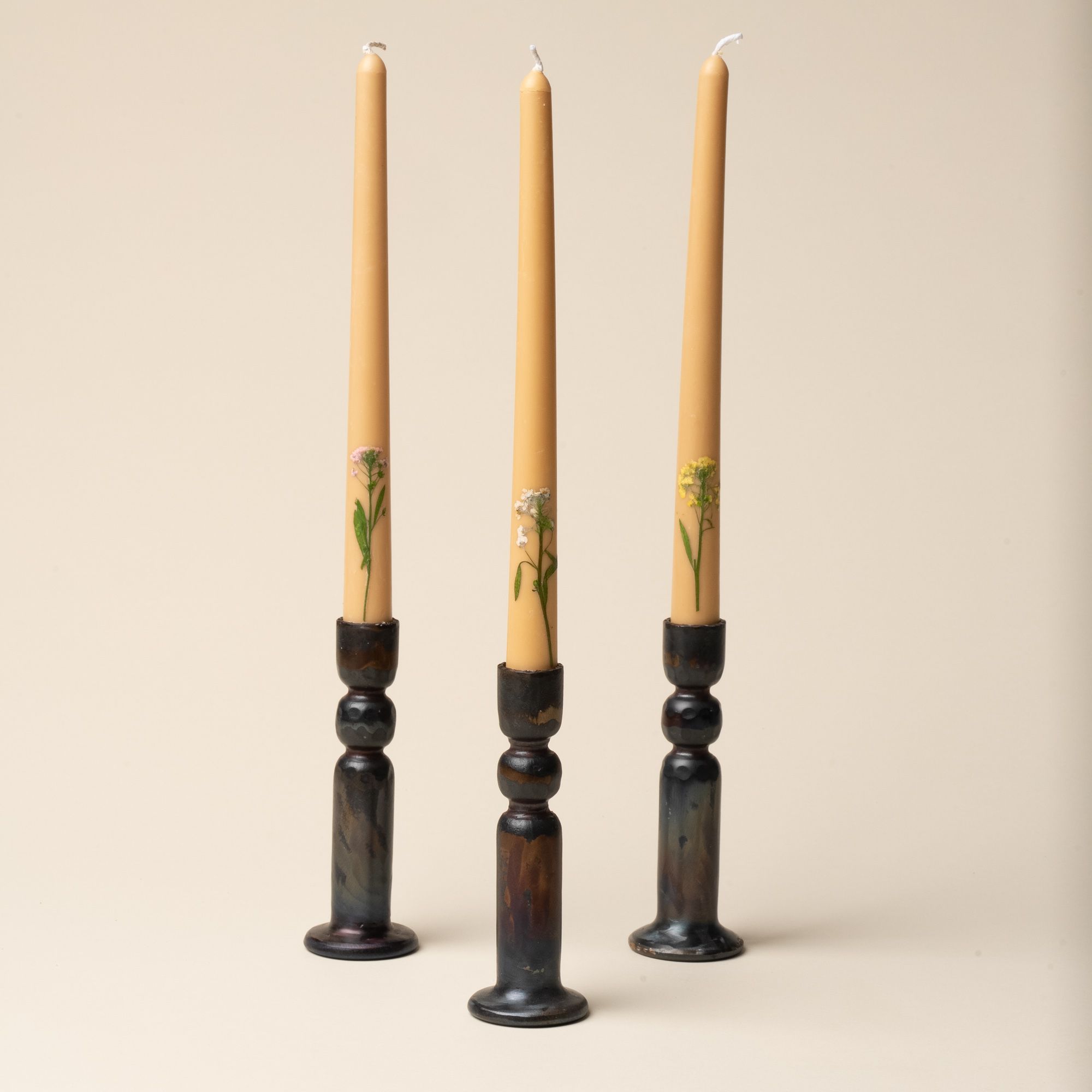 3 taper candles with pressed flowers on the bottom, all 3 sitting in a brutalist-styled iron candleholder.