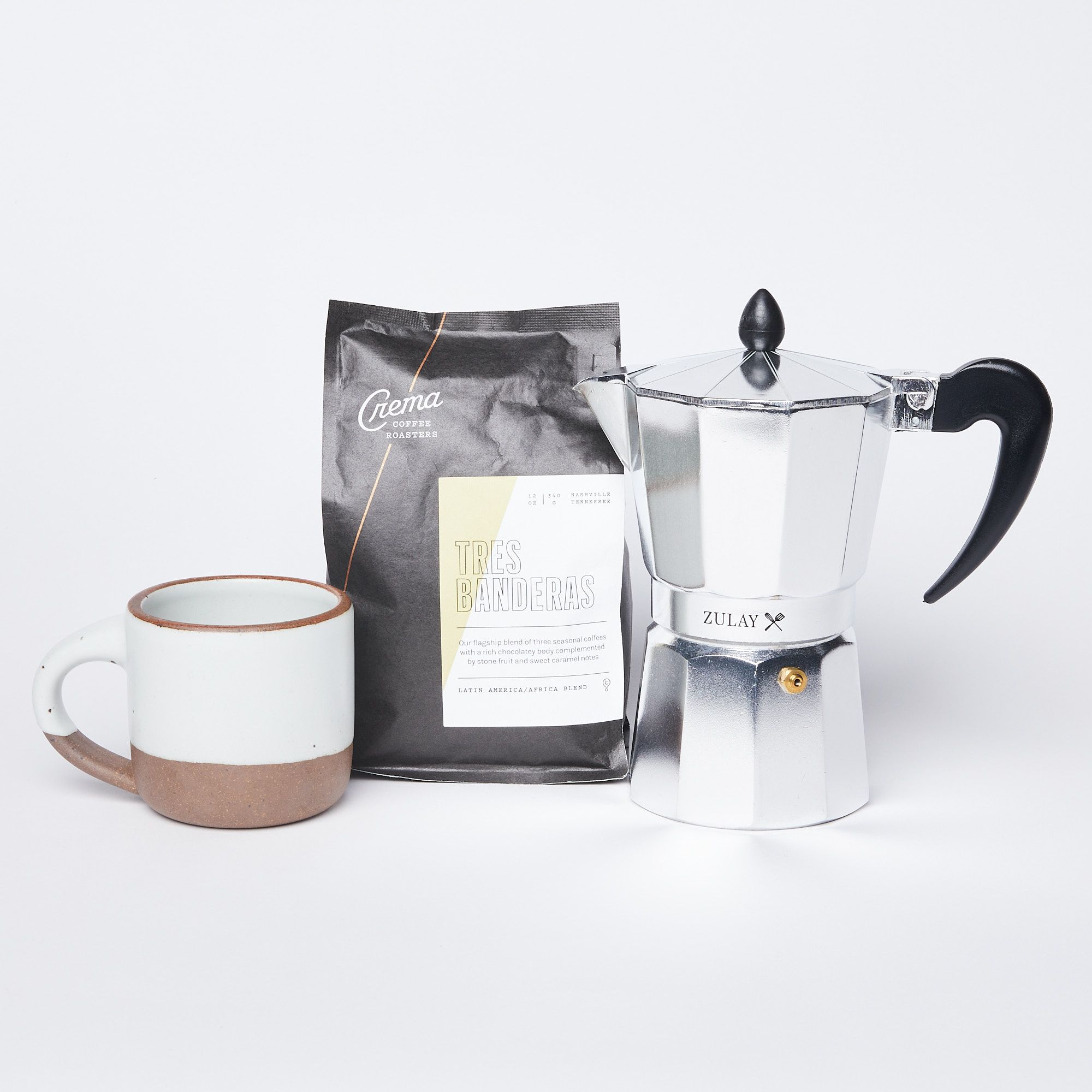 Small eggshell mug next to a black bag of coffee and a silver espresso maker