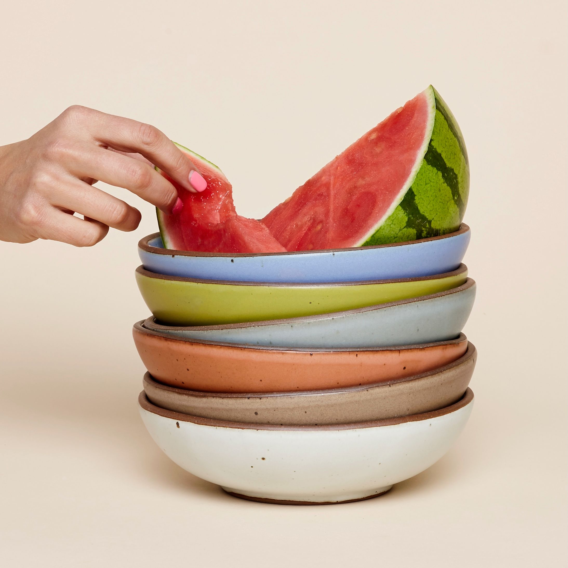 A hand reaches for a slice of watermelon sitting on a stack of colorful ceramic shallow bowls.