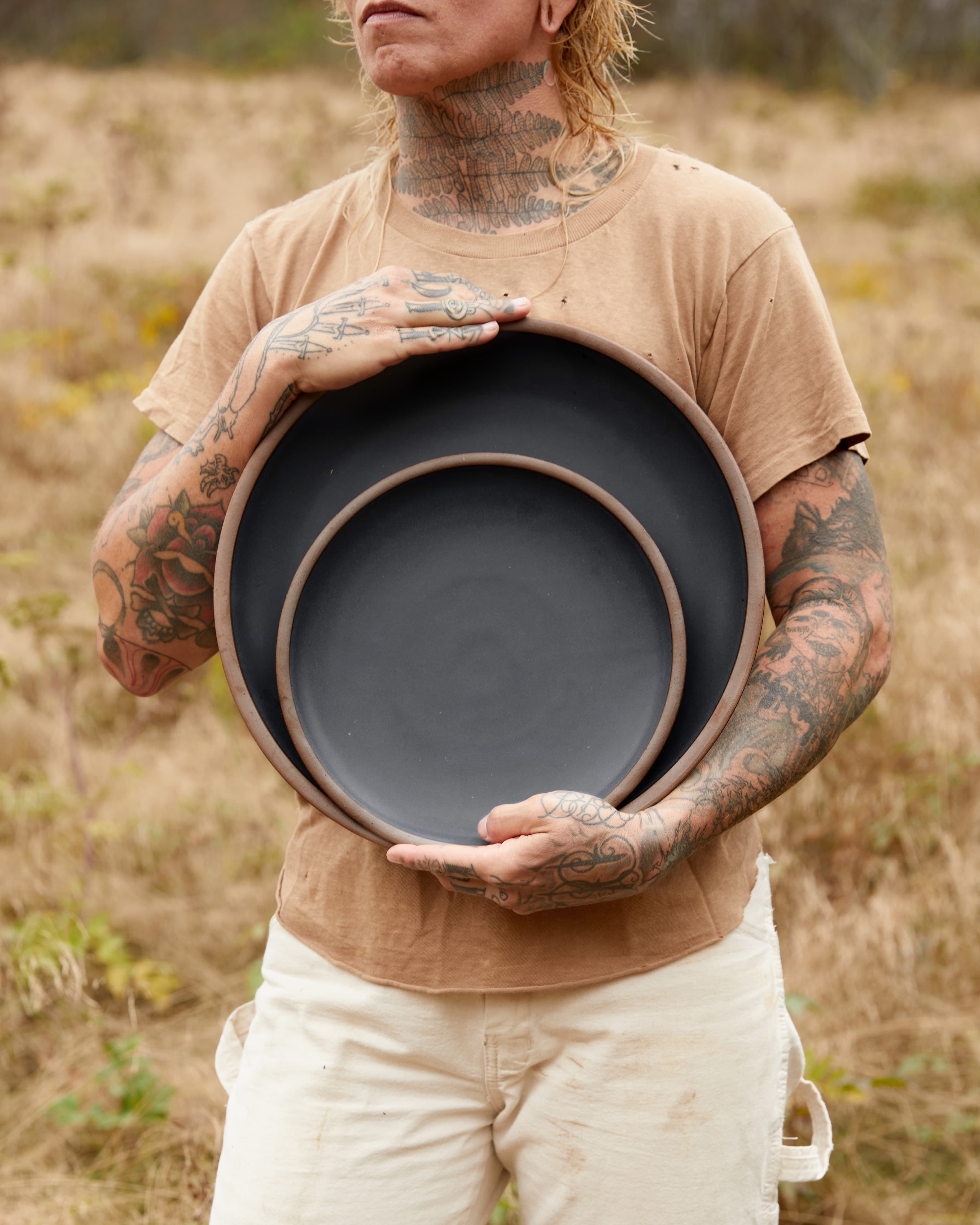 A person holds 2 large black ceramic plates outside with a field behind them.