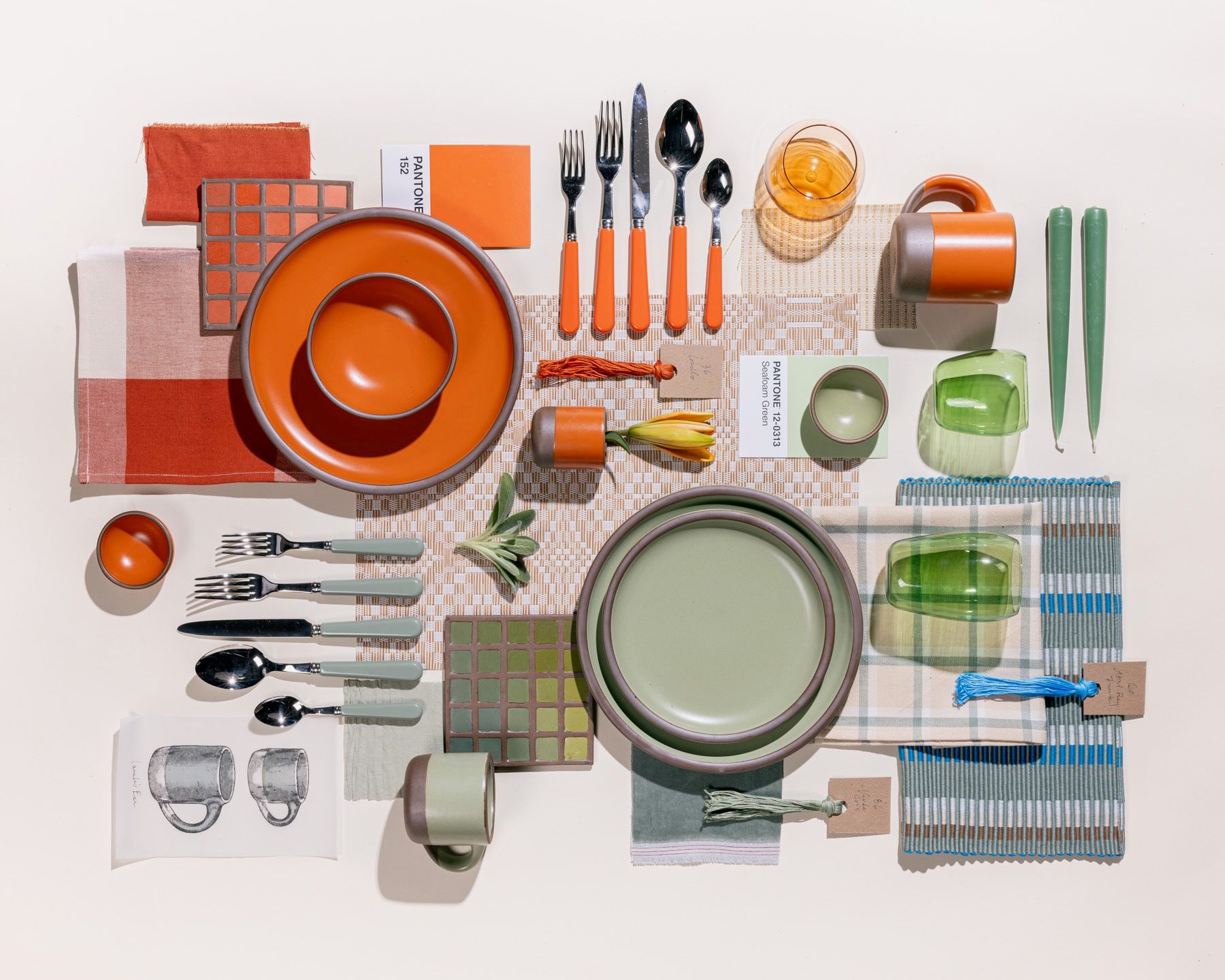 An overhead view of an artistic flat lay of ceramic plates in sage green and bold orange colors, surrounded by pantone cards, textile samples, flatware, glassware all in matching colors.