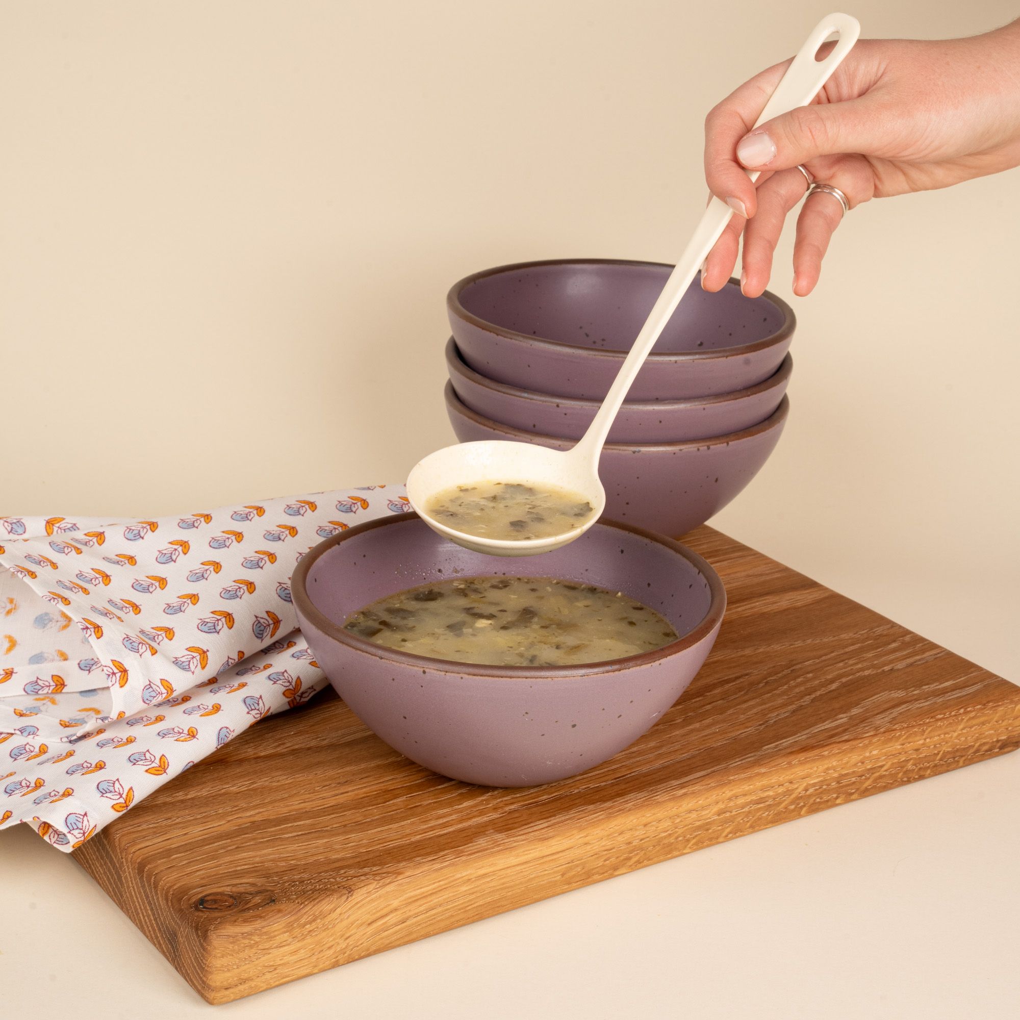 A hand holds a A hand holds a white enamel ladle scooping soup into a soft purple bowl near a napkin and stack of bowls.