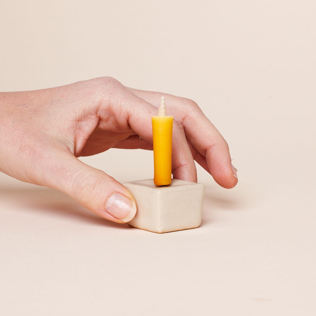 Two fingers of a hand hold a small square holder that has a short, thin yellow candle on it