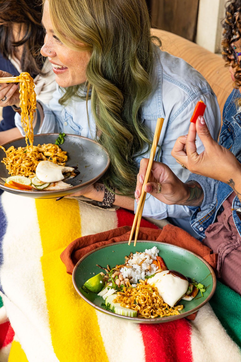 A woman with long hair hunched over a bowl of ramen smiling and holding chopsticks with ramen up to her mouth, about to take a bite