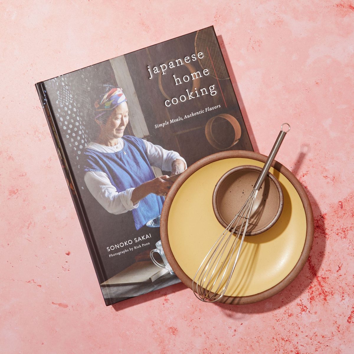 A plate and mini whisk rest on a a cookbook. The book's front cover is a photo of an older Japanese woman cooking with a title that reads "Japanese Home Cooking"