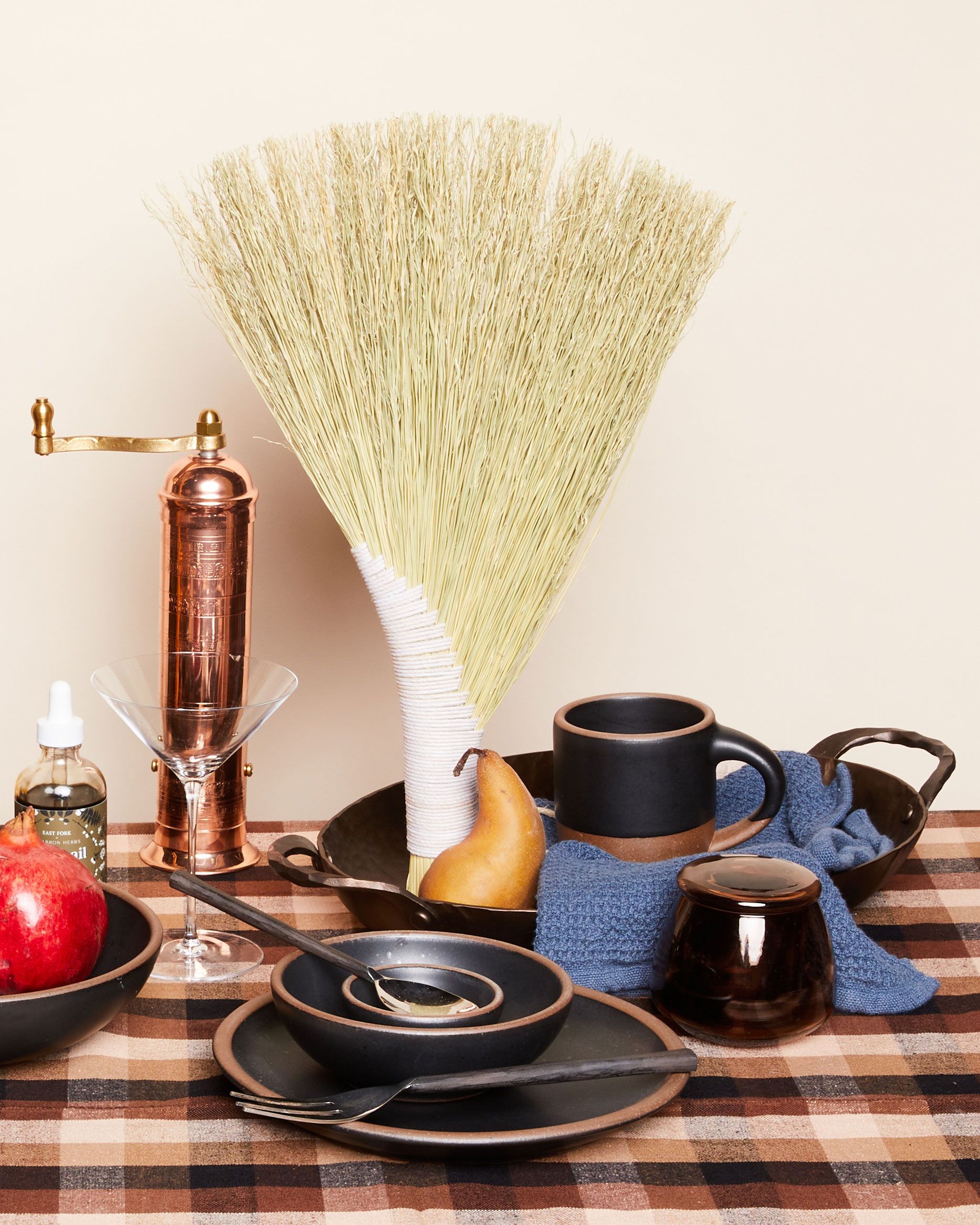 A table with a gingham tablecloth covered in black pottery, a pepper grinder, a martini glass, a butter keeper, a hand broom, martini bitters, and fruits. 