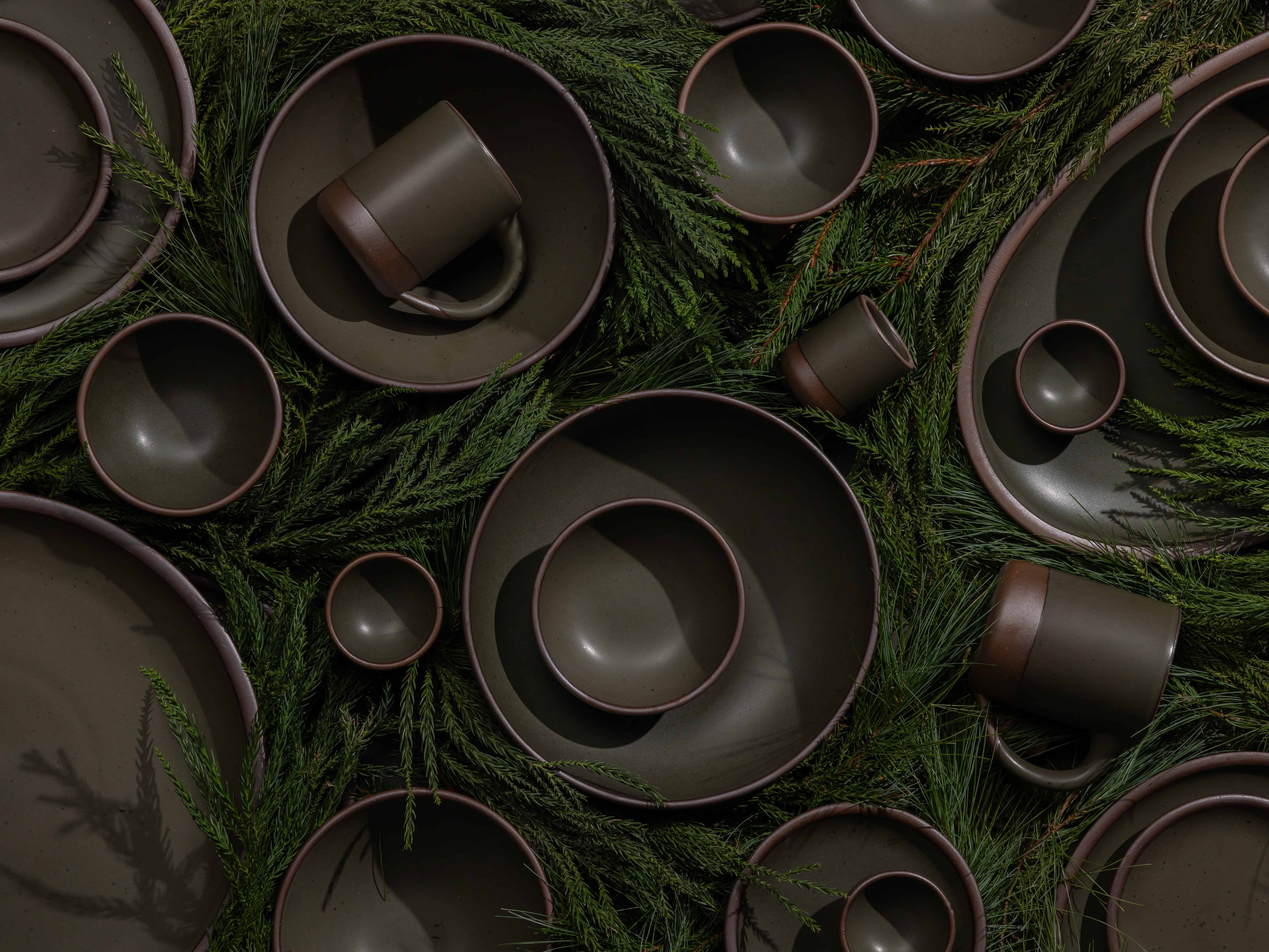 Ceramic plates, bowls, and mugs in a forest green artfully laid out on a bed of pine leaves.