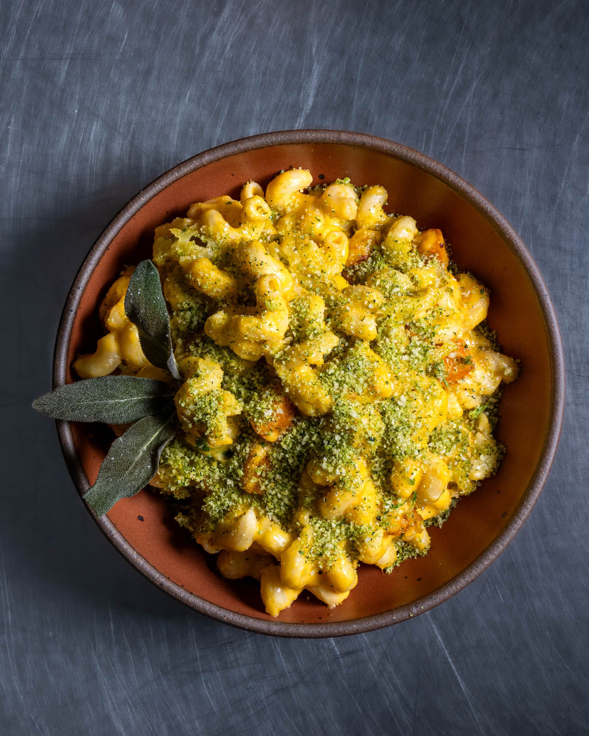 A ceramic bowl in a terracotta color is filled with a gourmet mac and cheese dish with garnish