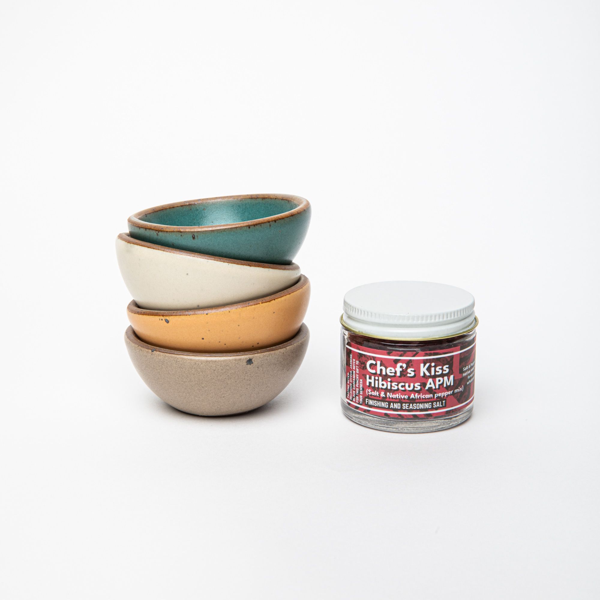 A stack of bitty bowls in turquoise, cream, soft peachy yellow, and light earthy brown - next to a small jar with a white top and pink label that reads "Chef's Kiss Hibiscus APM"