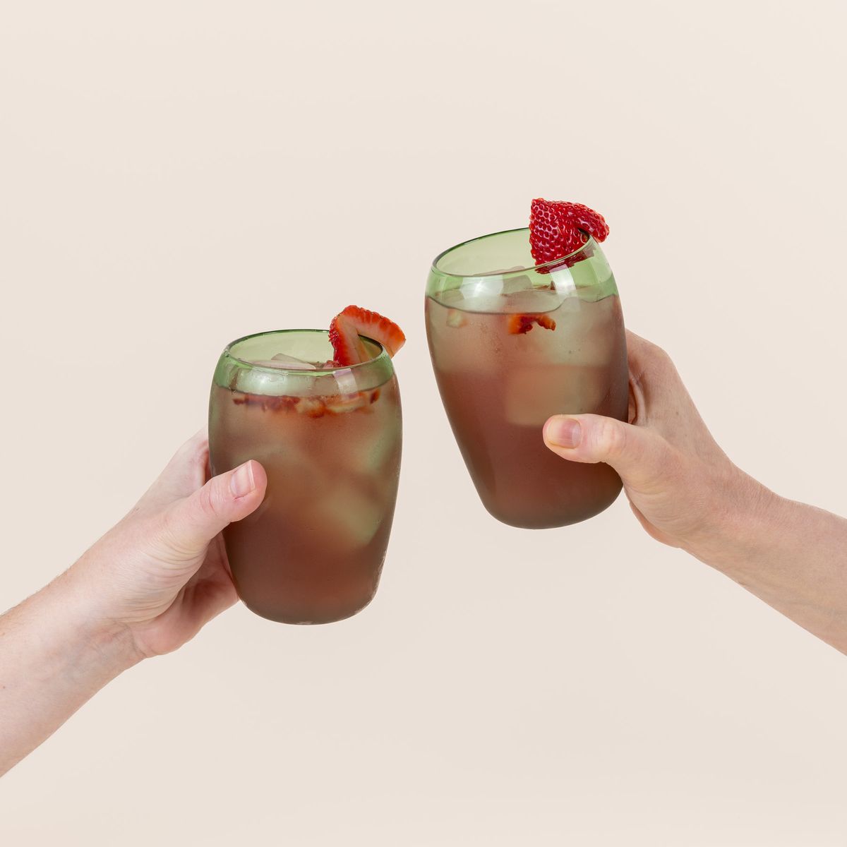 Hands are holding two tall light green glass tumblers with a slight curve inward on the top. The glasses are filled with a red cocktail with sliced strawberries on the rim.