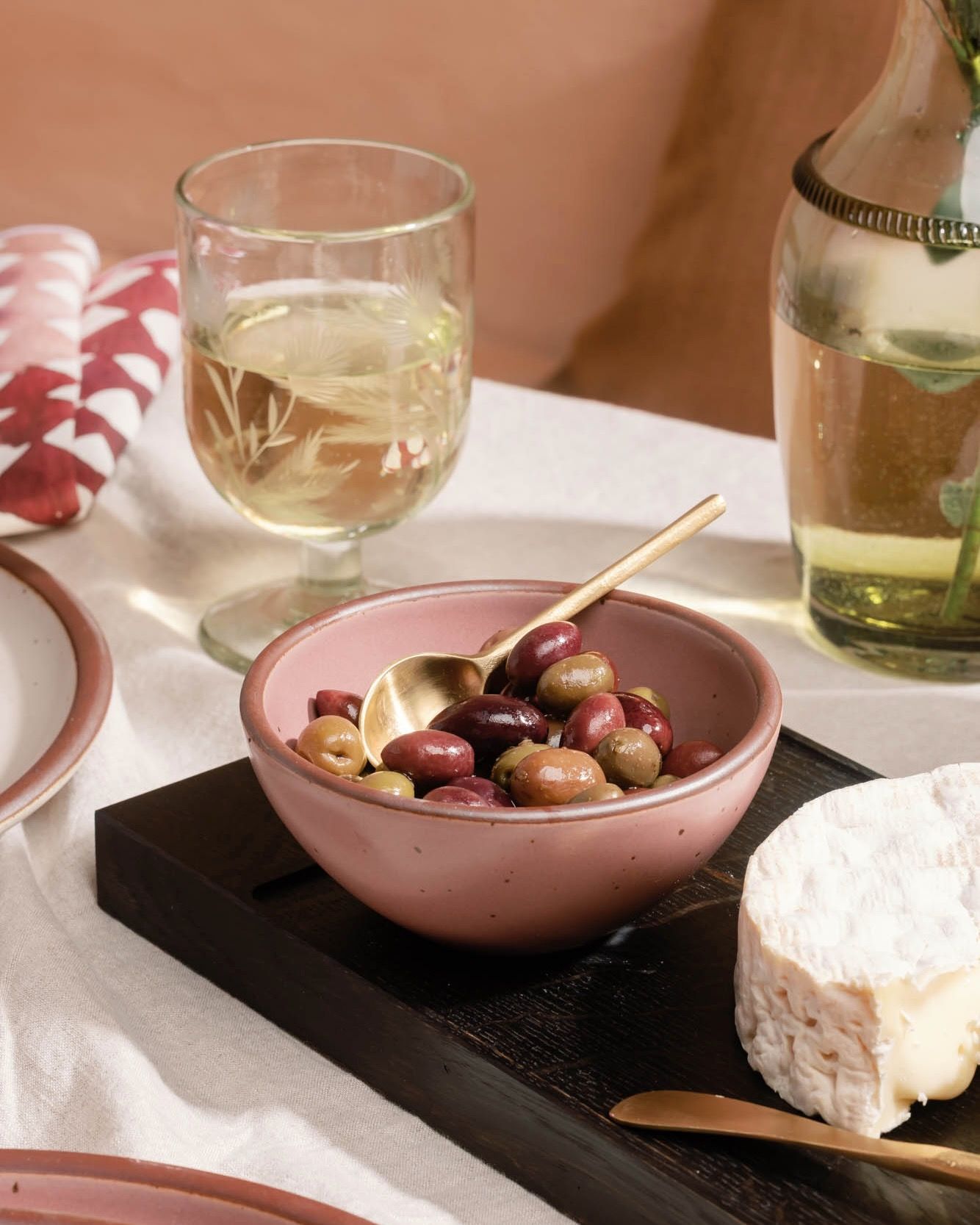 Small pink ceramic bowl filled with olives and a brass spoon, set on a wooden serving board with wine glasses nearby.