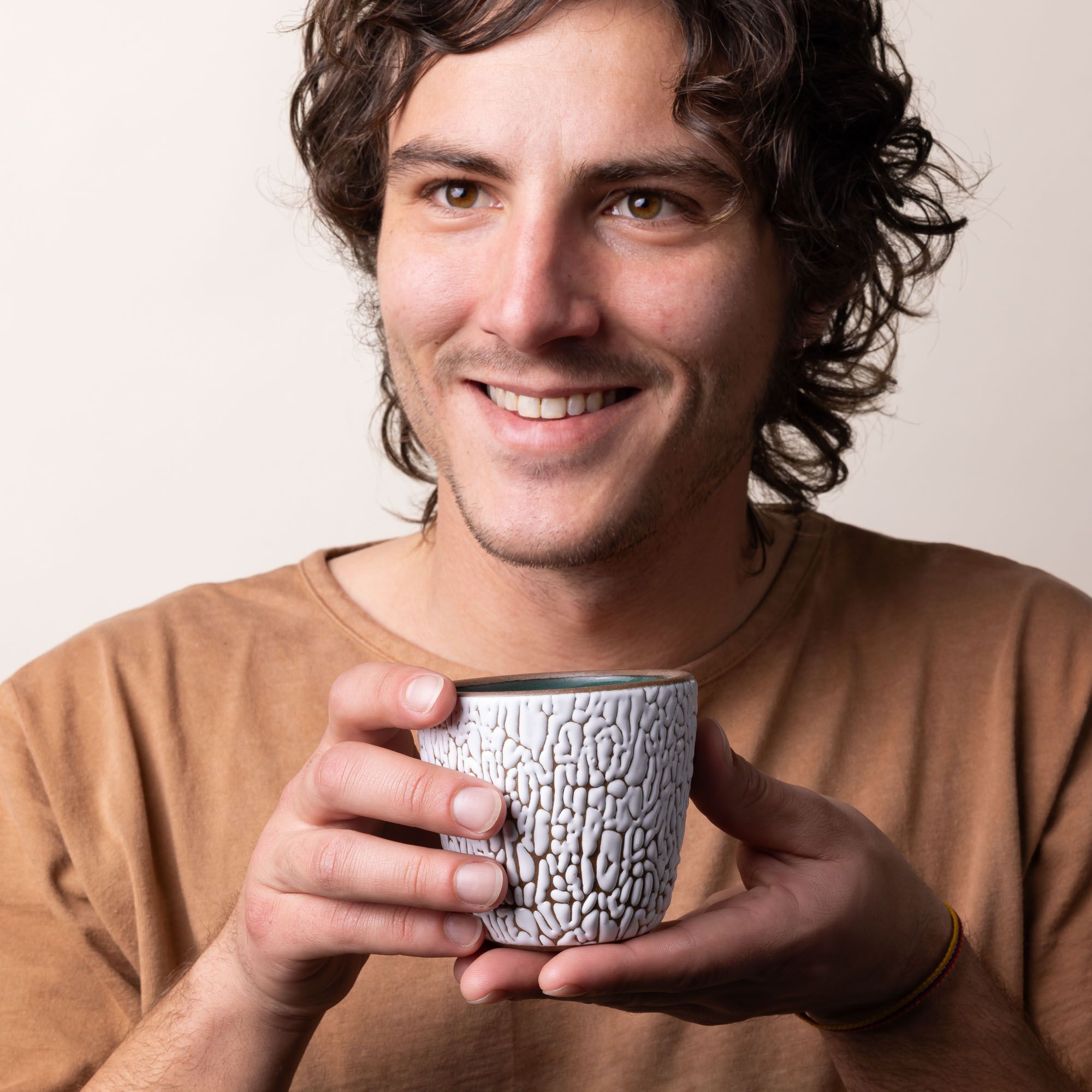 A person is smiling and holding a small ceramic cup with cracked texture and the interior being dark teal.
