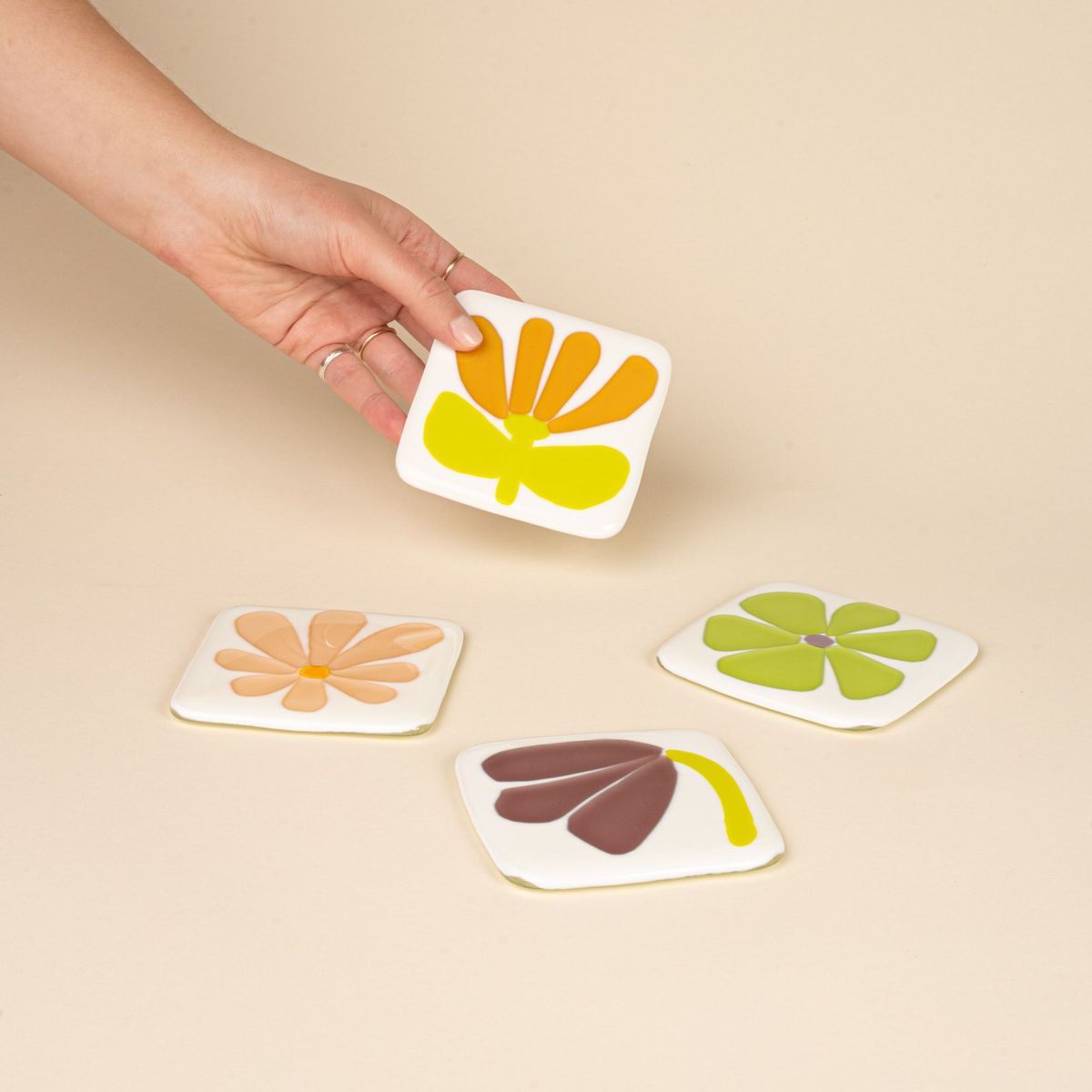 A hand picks up one of four coasters featuring simple painterly flower shapes in peach, amber, purple, and neon green.