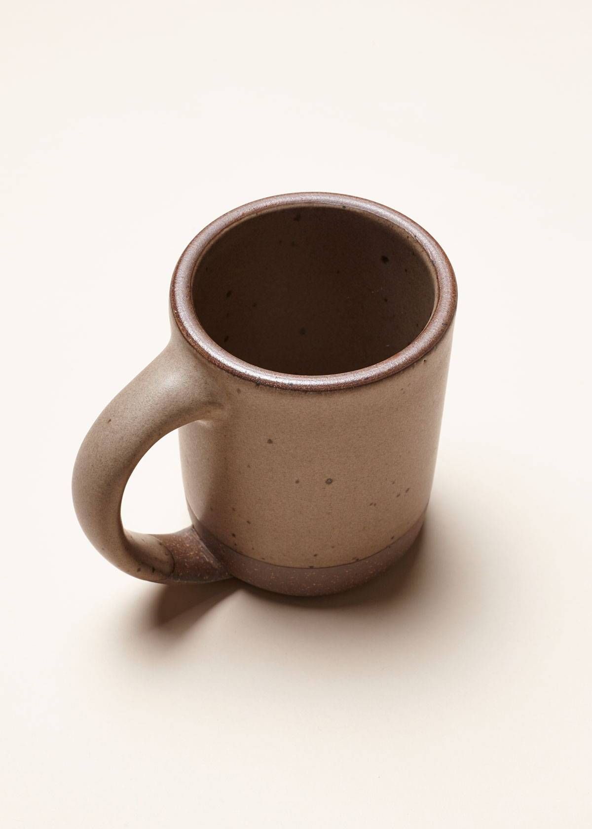 A medium sized ceramic mug with handle in a warm pale brown color featuring iron speckles and unglazed rim and bottom base.