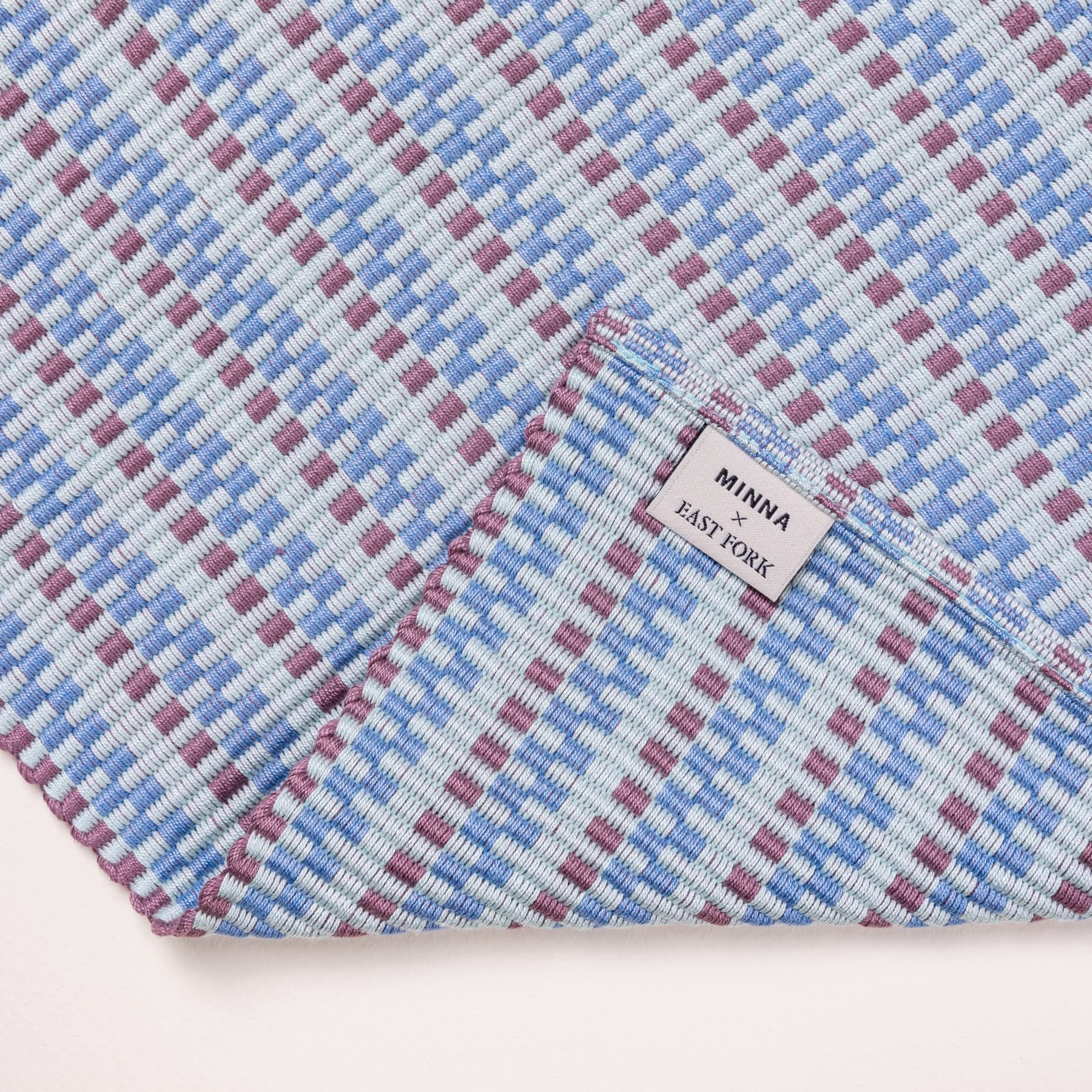 A corner of a woven placemat with thin horizontal striped tonal soft blue and purple colors. A tag reads 'Minna x East Fork'