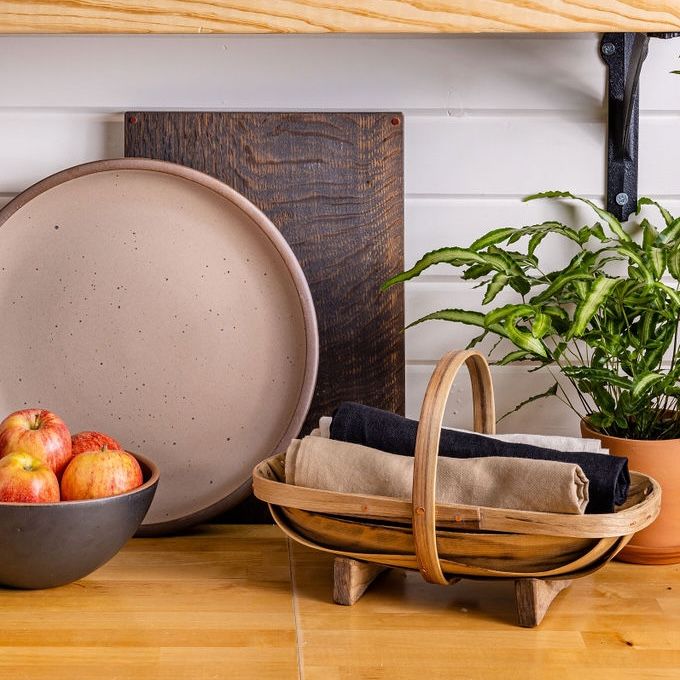 On a kitchen counter is a wooden trug with rolled up napkins in it, a large serving platter, a cutting board, and bowl filled with apples.
