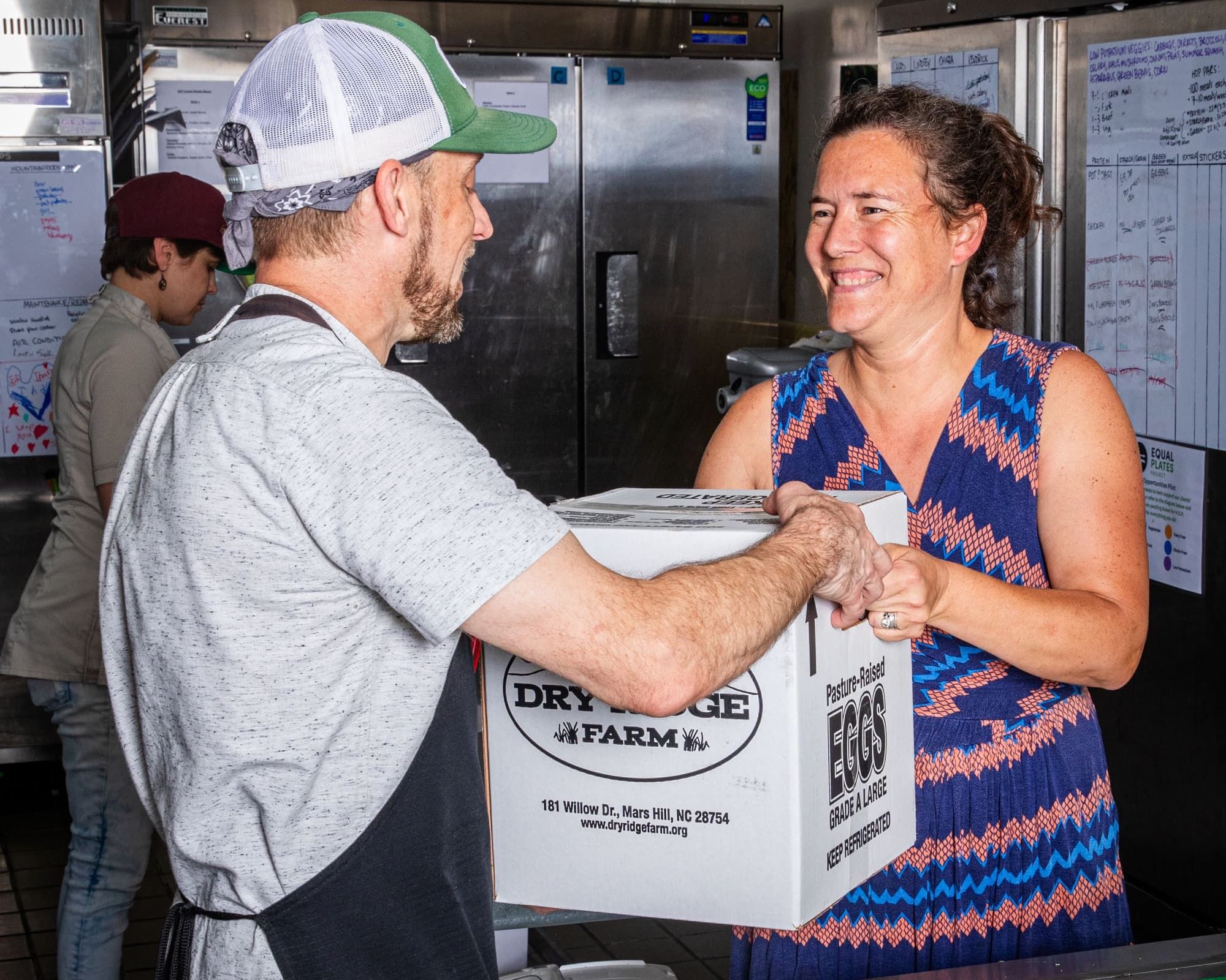 In a kitchen, a person hands a box of eggs to another person and is smiling.