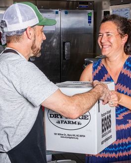 In a kitchen, a person hands a box of eggs to another person and is smiling.