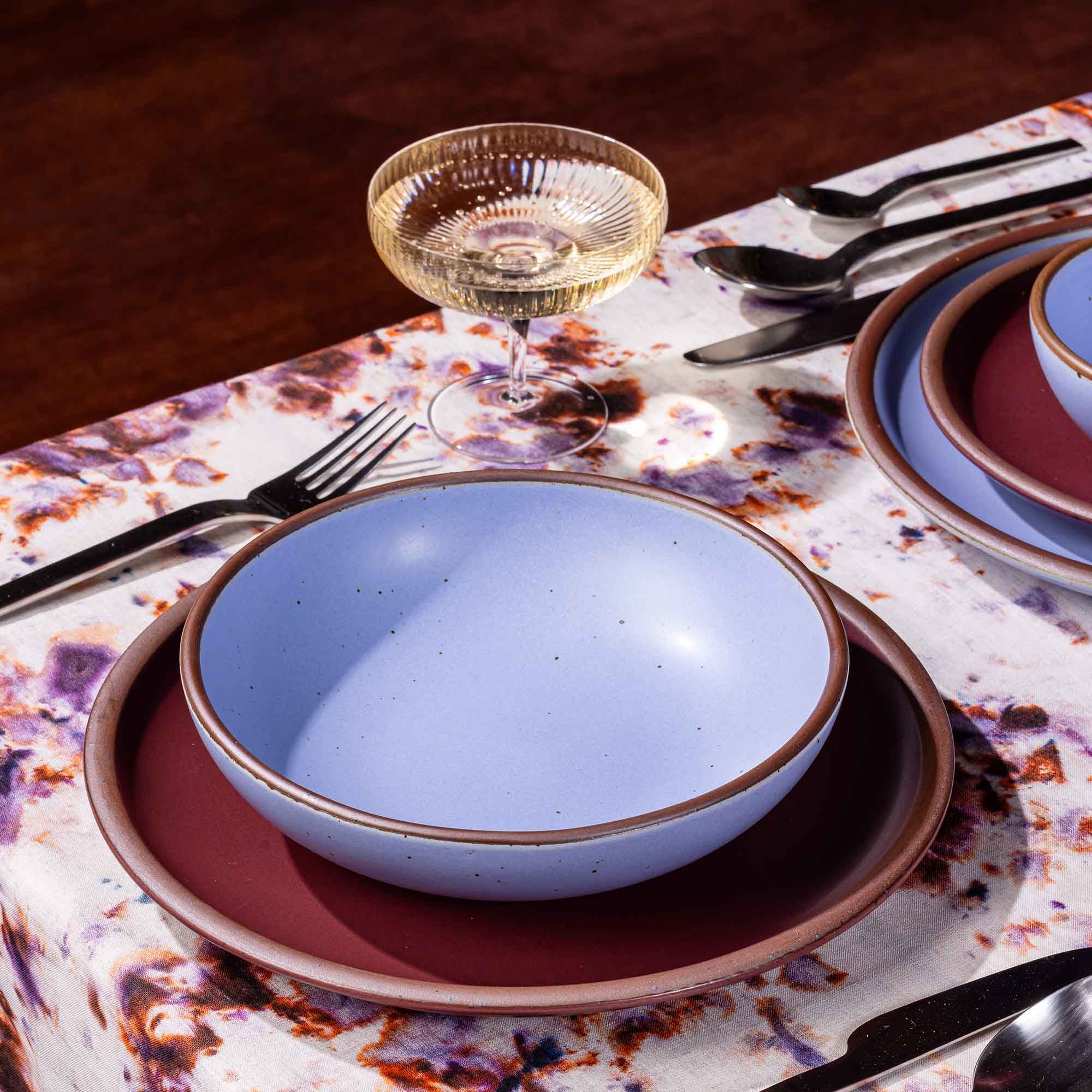 A table setting featuring marbled textiles, a coupe glass, and a plate and shallow bowl in plum and periwinkle colors