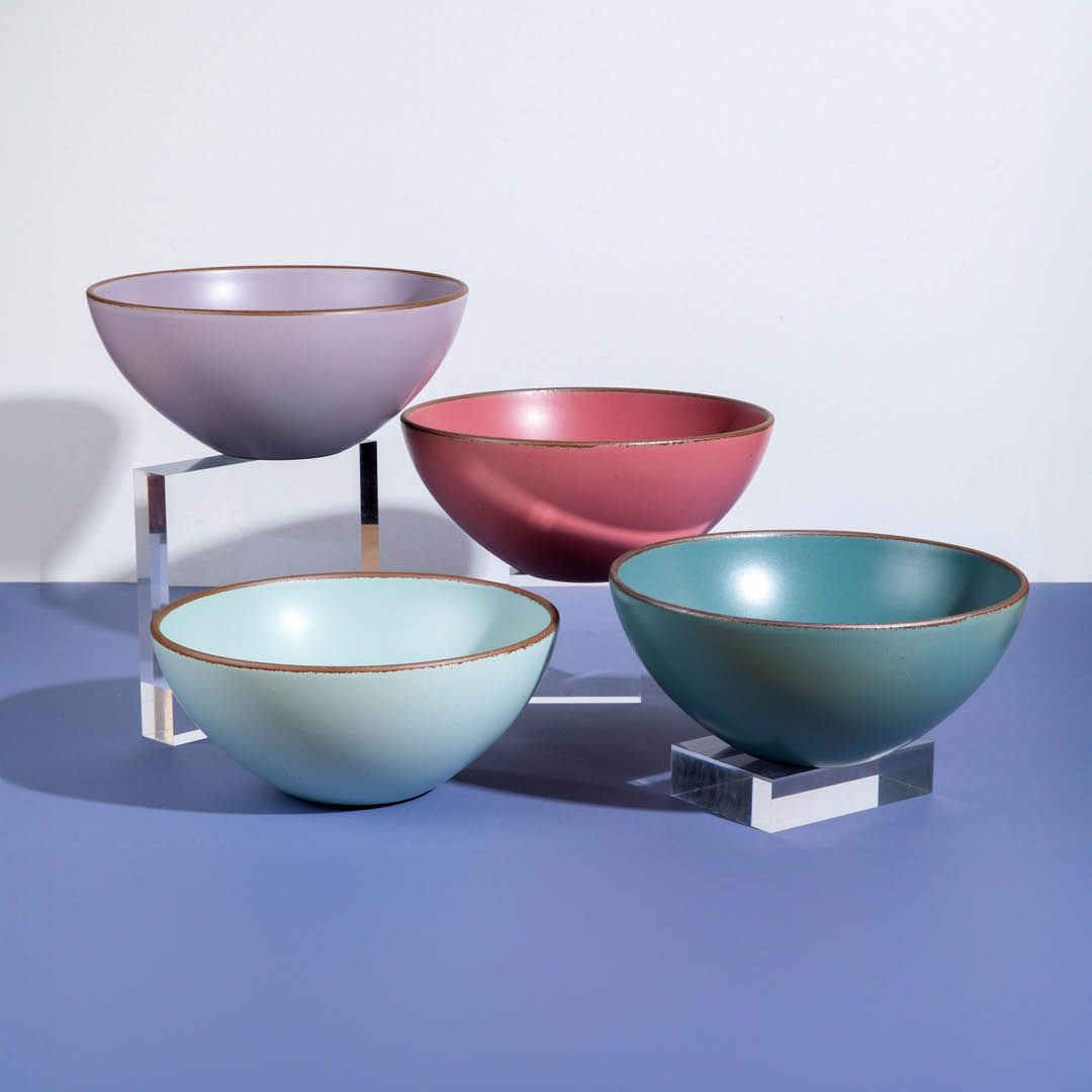 4 Ceramic large mixing bowls in 4 different colors sit in a studio setting on acrylic risers. The colors of the bowls are soft muted lilac, cool pink, baby blue, and turquoise.