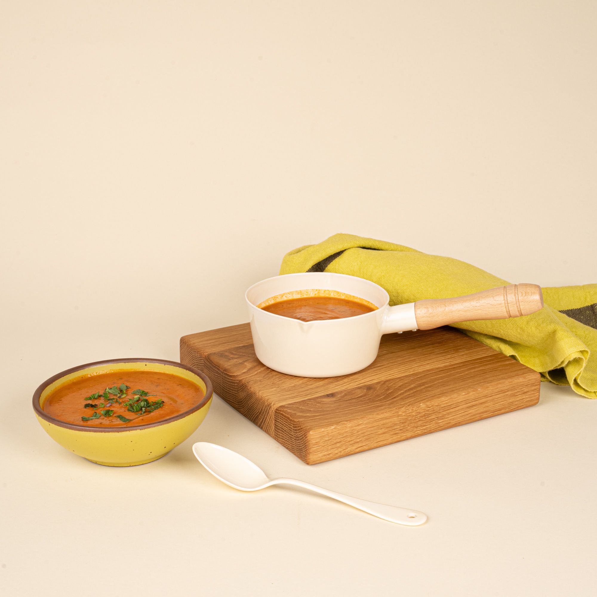 A white enamel pot with a wooden handle filled with soup next to a yellow soup bowl