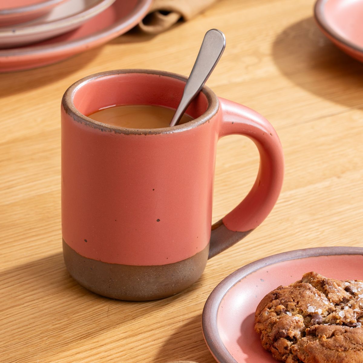 Watermelon pink ceramic mug filled with coffee and a spoon, next to a matching plate with a chocolate chip cookie on a wood table.