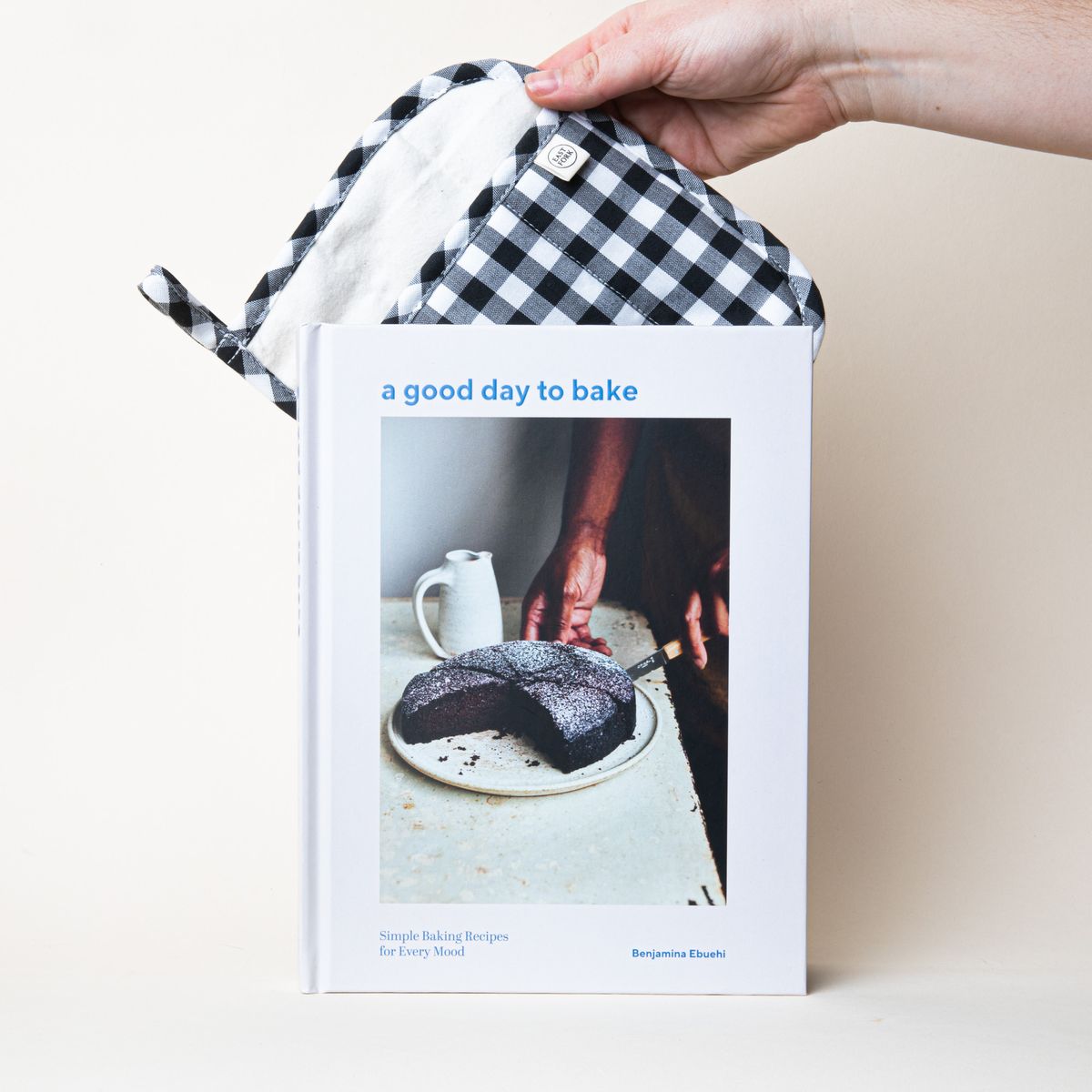 A hand holding a pot holder behind a book with a white cover and a large photo of a person cutting a cake with a title that reads "a good day to bake"