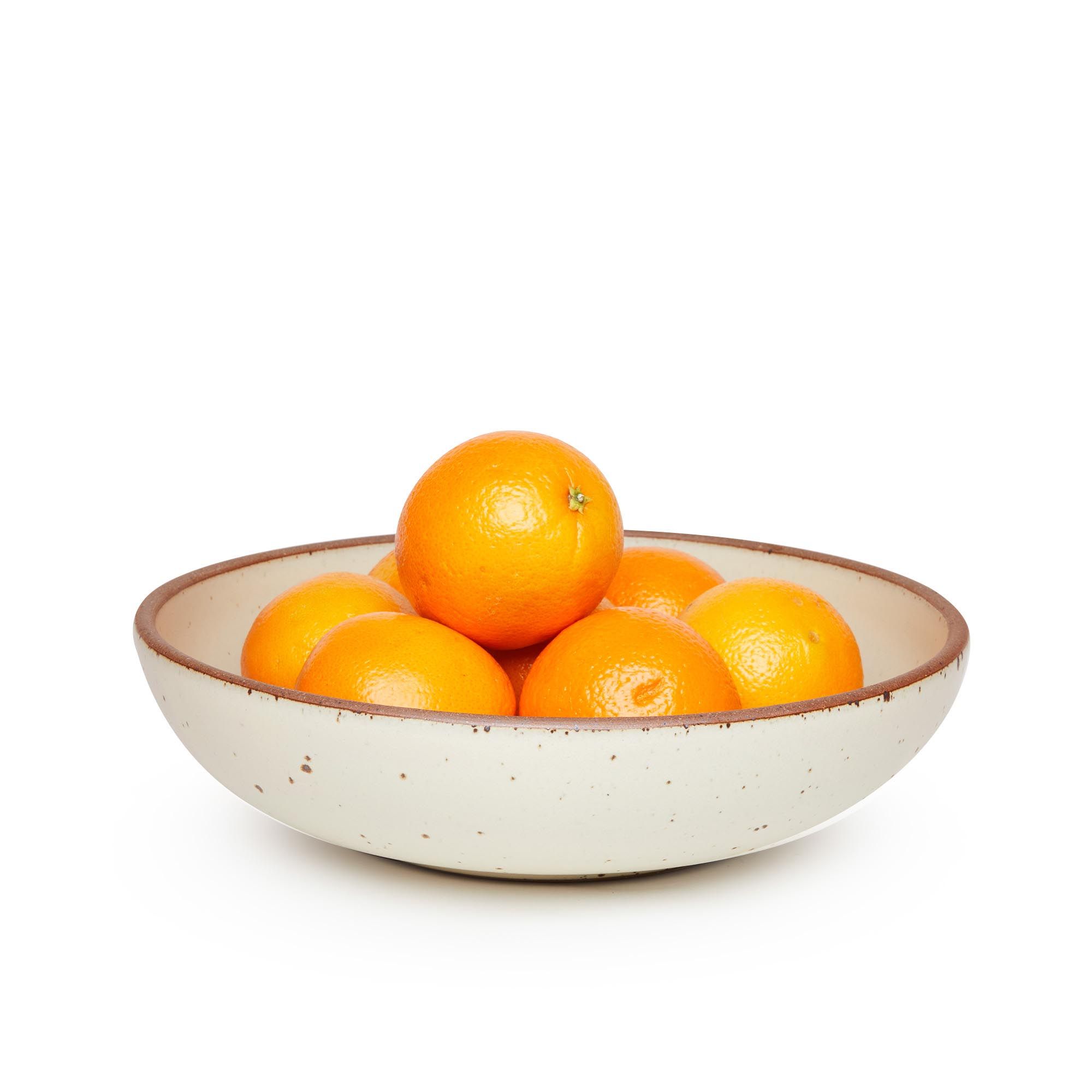 A large shallow serving ceramic bowl in a warm, tan-toned, off-white color featuring iron speckles and an unglazed rim, filled with oranges