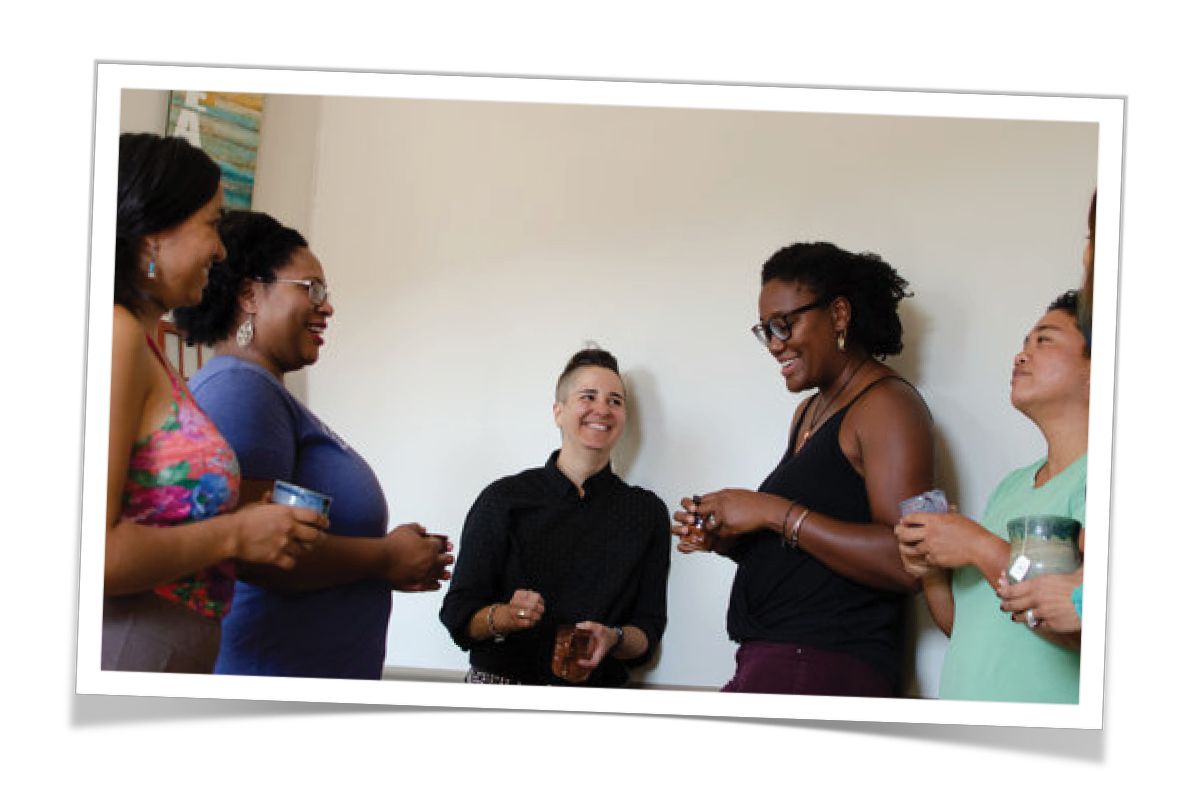 5 people gathered around in a standing circle holding cups and chatting and smiling