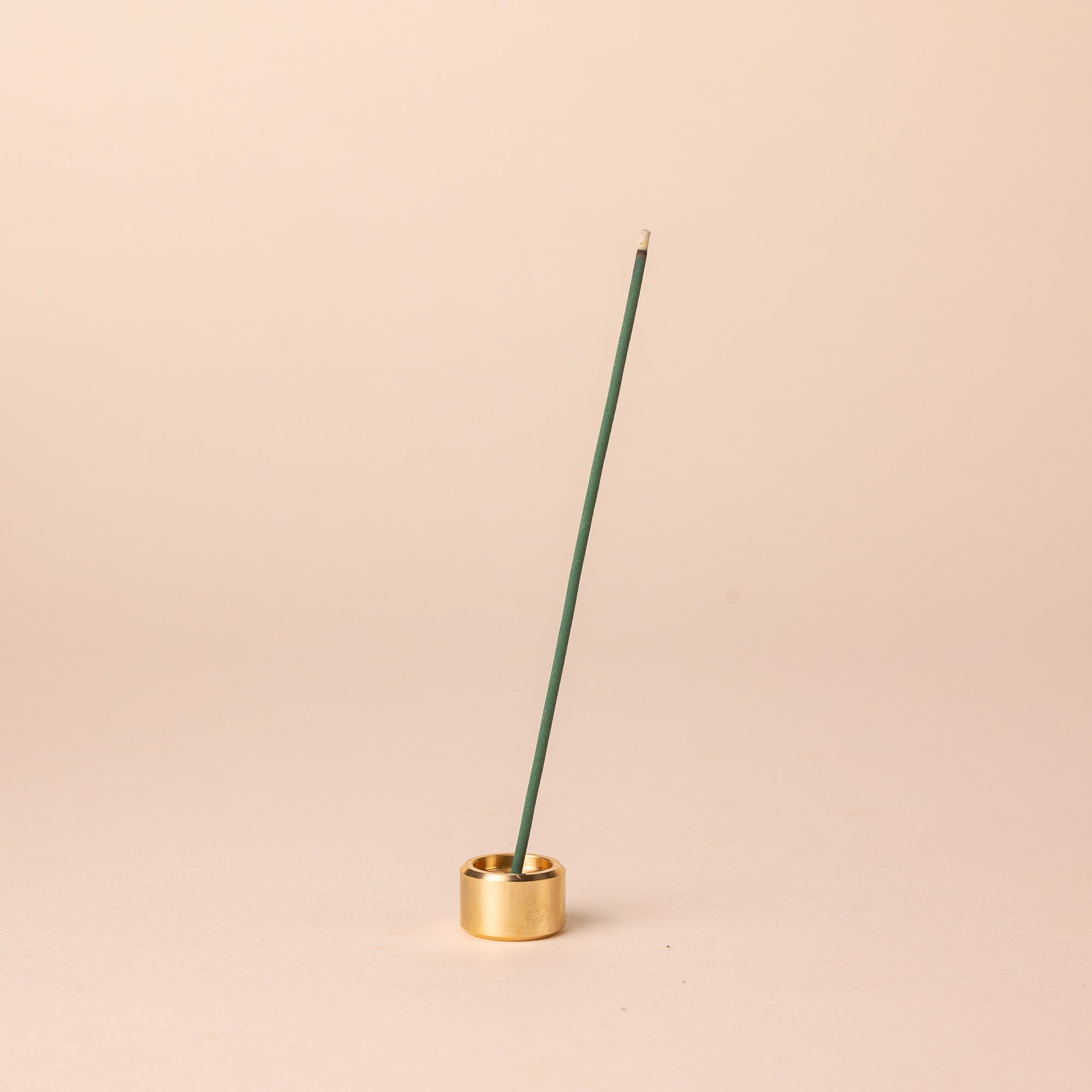 A short small brass cylinder incense holder with a teal incense stick sitting on top.