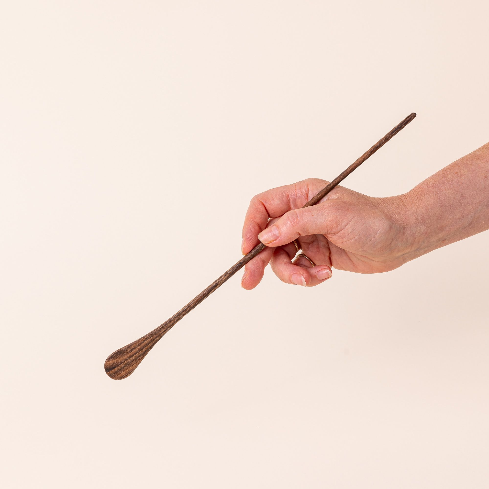 A hand holds a long thin walnut cocktail stirrer with a small spoon-like shape on the end