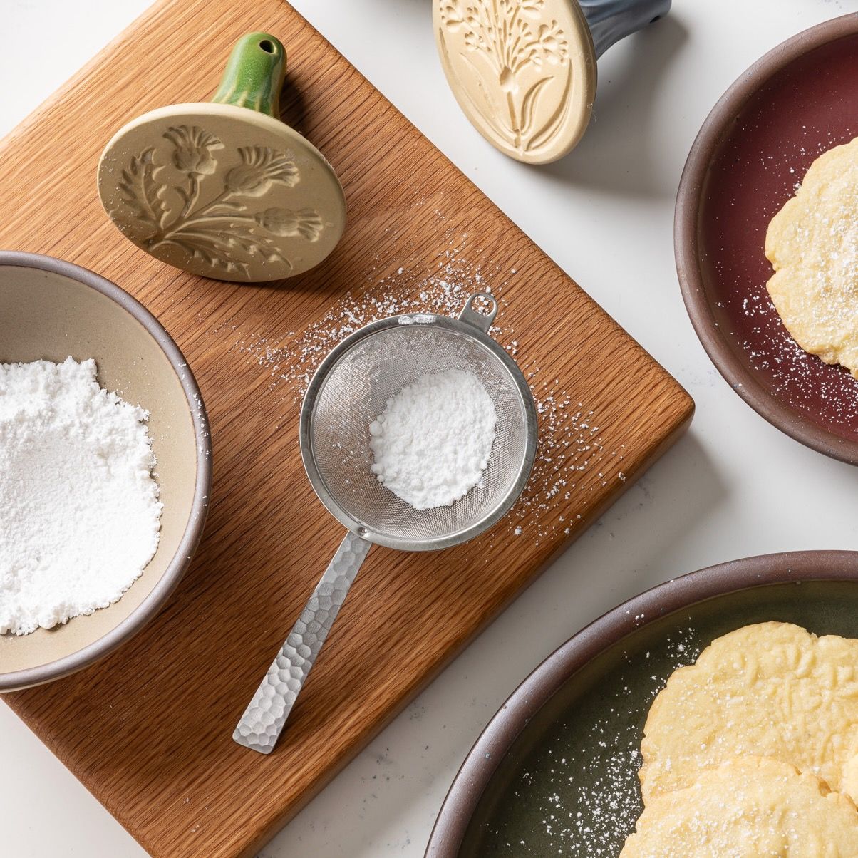 Stainless steel half dome strainer with steel handle on a cutting board with powdered sugar for cookies.