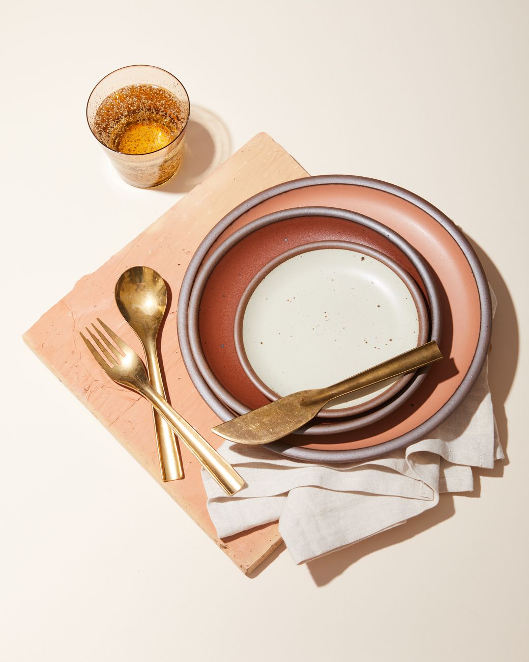 A table setting with three plates in a soft nude (Utah), a burnt red (Amaro), and an off-white (Panna Cotta). The setting also shows brass flatware, a natural-white napkin, and an orange water glass.