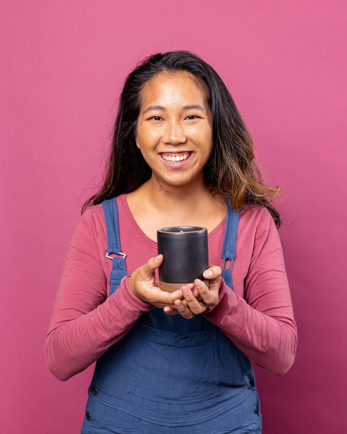 Kate is smiling against a pink backdrop and holding a large candle in a black ceramic vessel