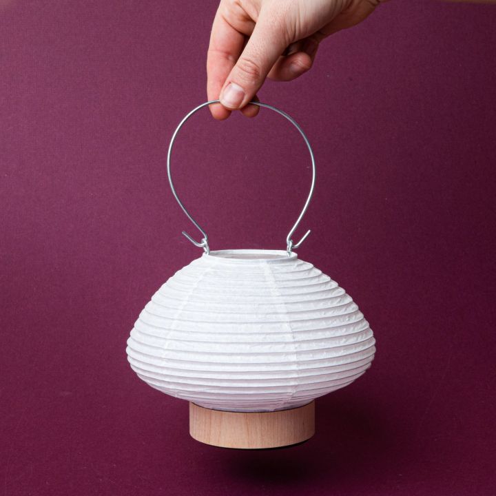 A hand holding a white paper lantern in a short horizontal oval shape with a wood base and a thin metal handle
