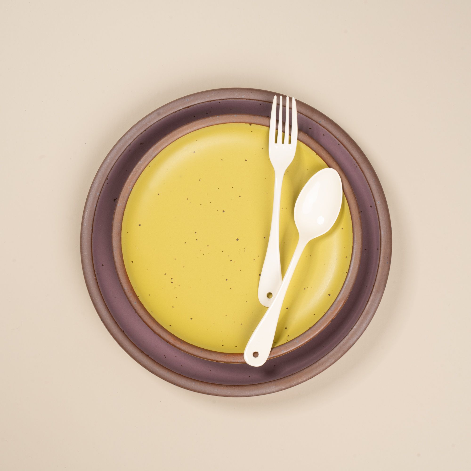 A white enamel fork and spoon sitting on a stacked zesty yellow and soft purple plate.