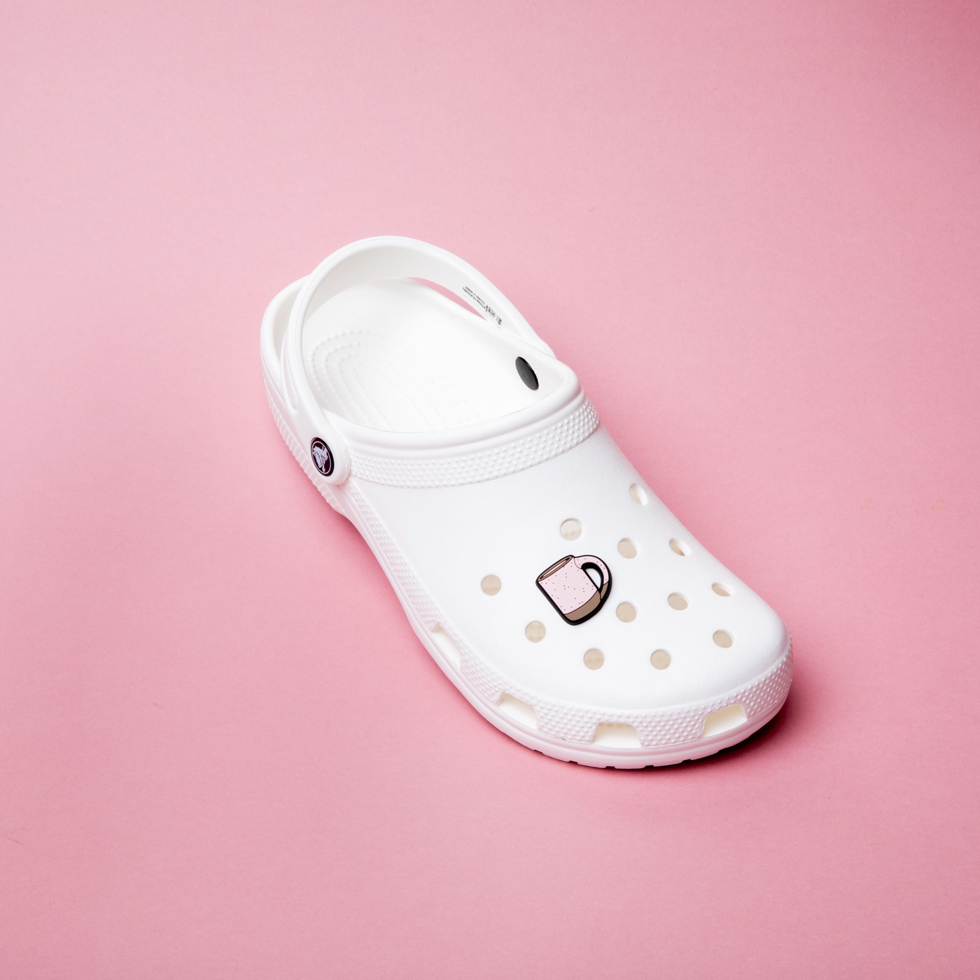 A white plastic clog made by the brand Crocs with a soft light pink charm on the top that looks like an East Fork mug