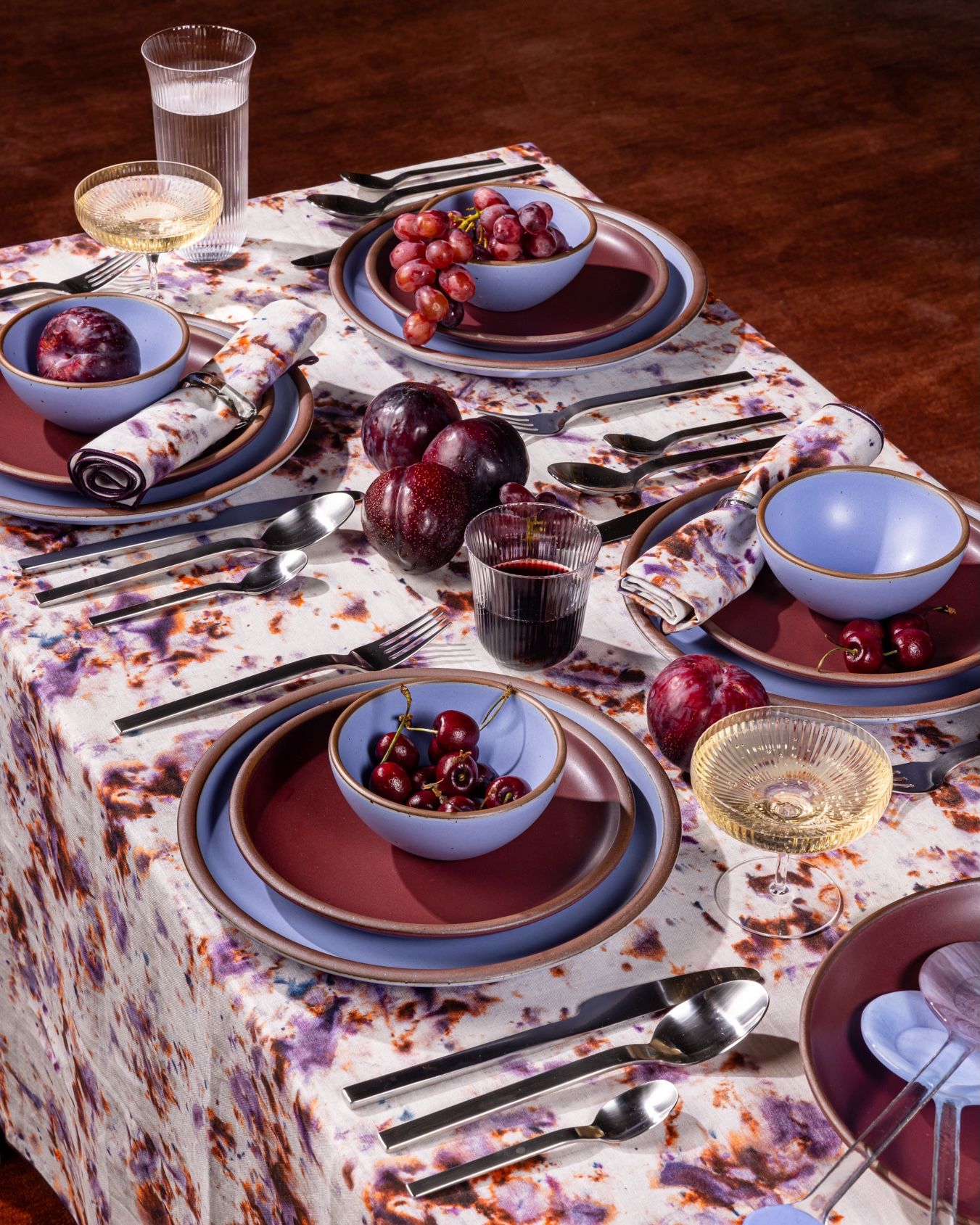A colorful tablescape with ceramic plates and bowls in periwinkle and plum, wine glasses, and silverware.