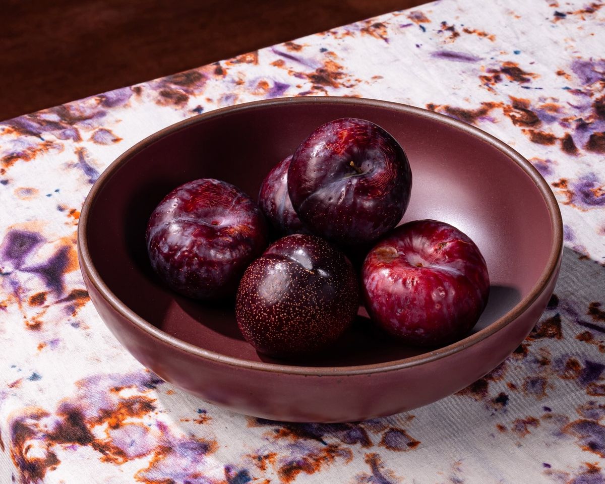 Plums sit in a large shallow serving ceramic bowl in a plum color on a colorful marbled tablecloth