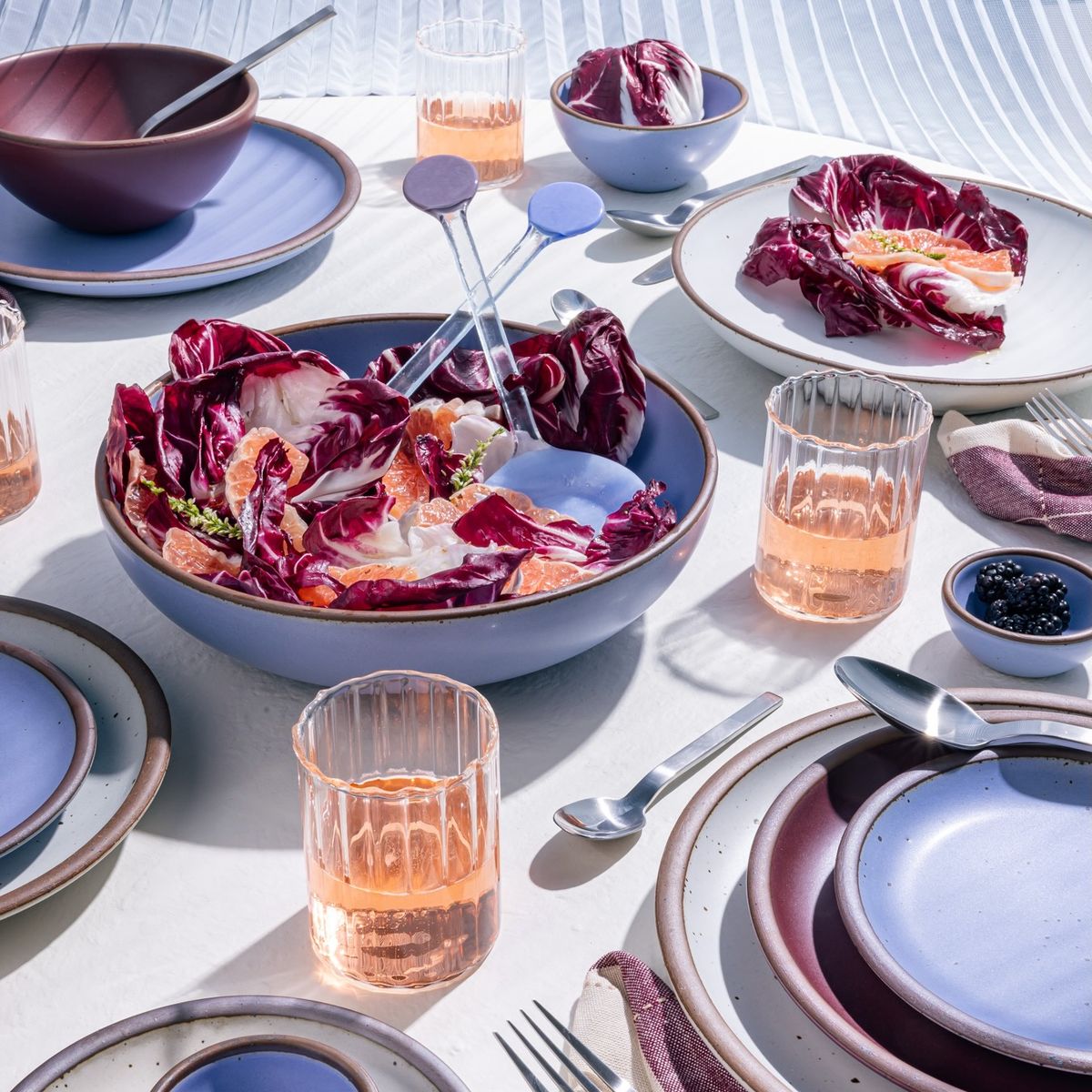 A table filled with ceramic dinnerware in periwinkle, plum, and cool white colors, along with silverware and fluted short glasses filled with rosé.