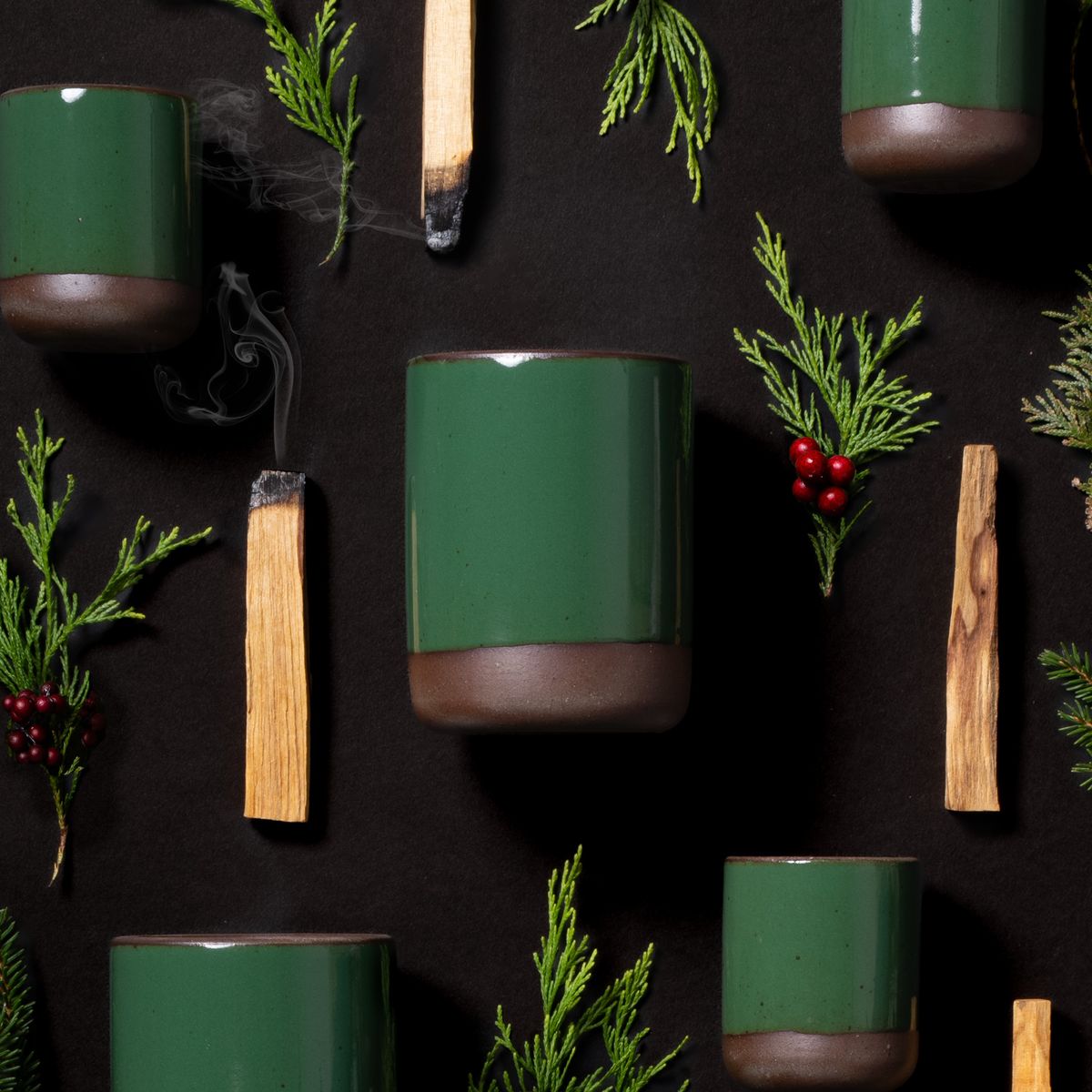 Glossy emerald green ceramic candles with unglazed bases arranged on black background with cedar sprigs, berries, and palo santo sticks.