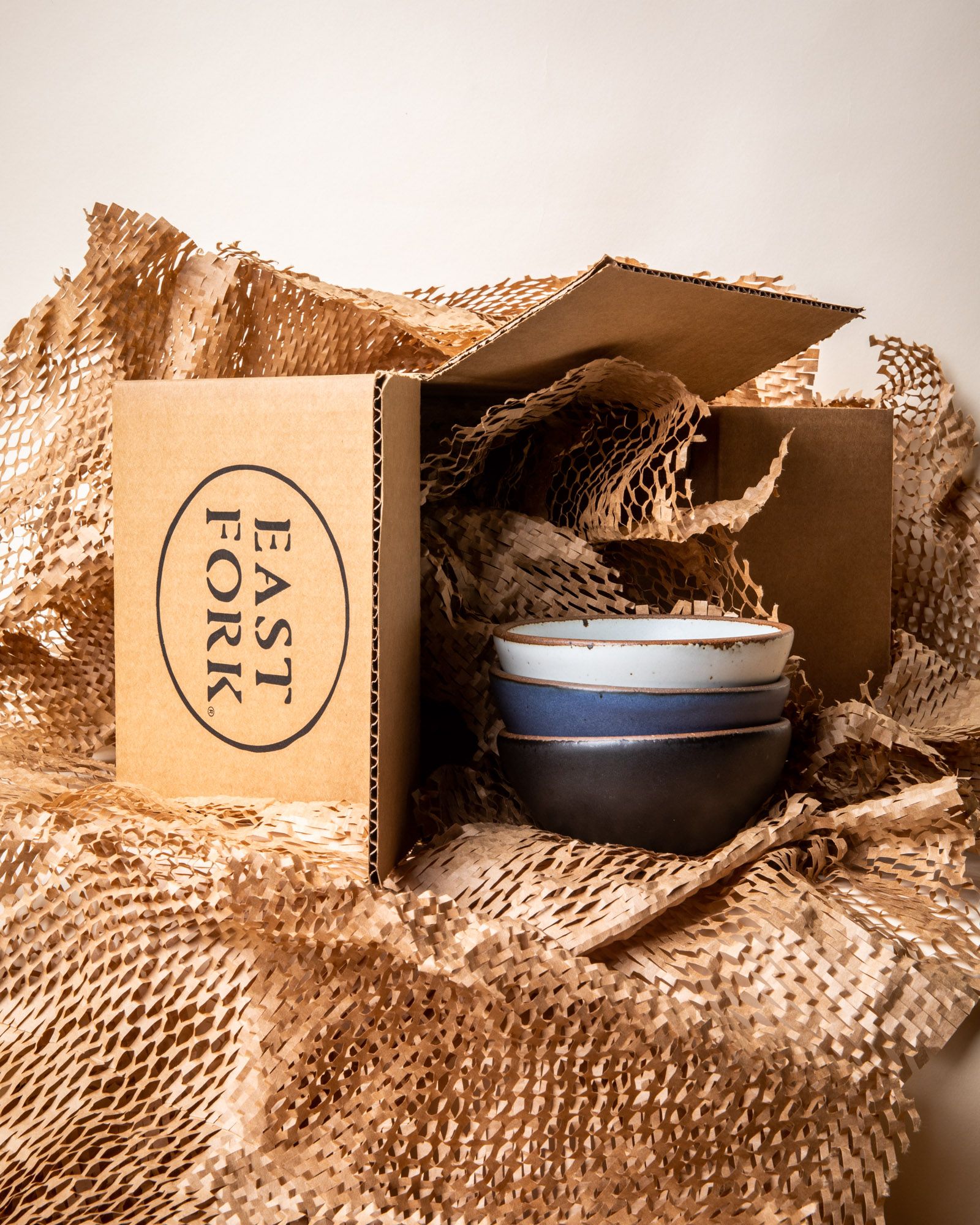 A kraft box that says "East Fork on its side with 3 ceramic bowls in white, blue, and black. All laying in geami paper packaging.